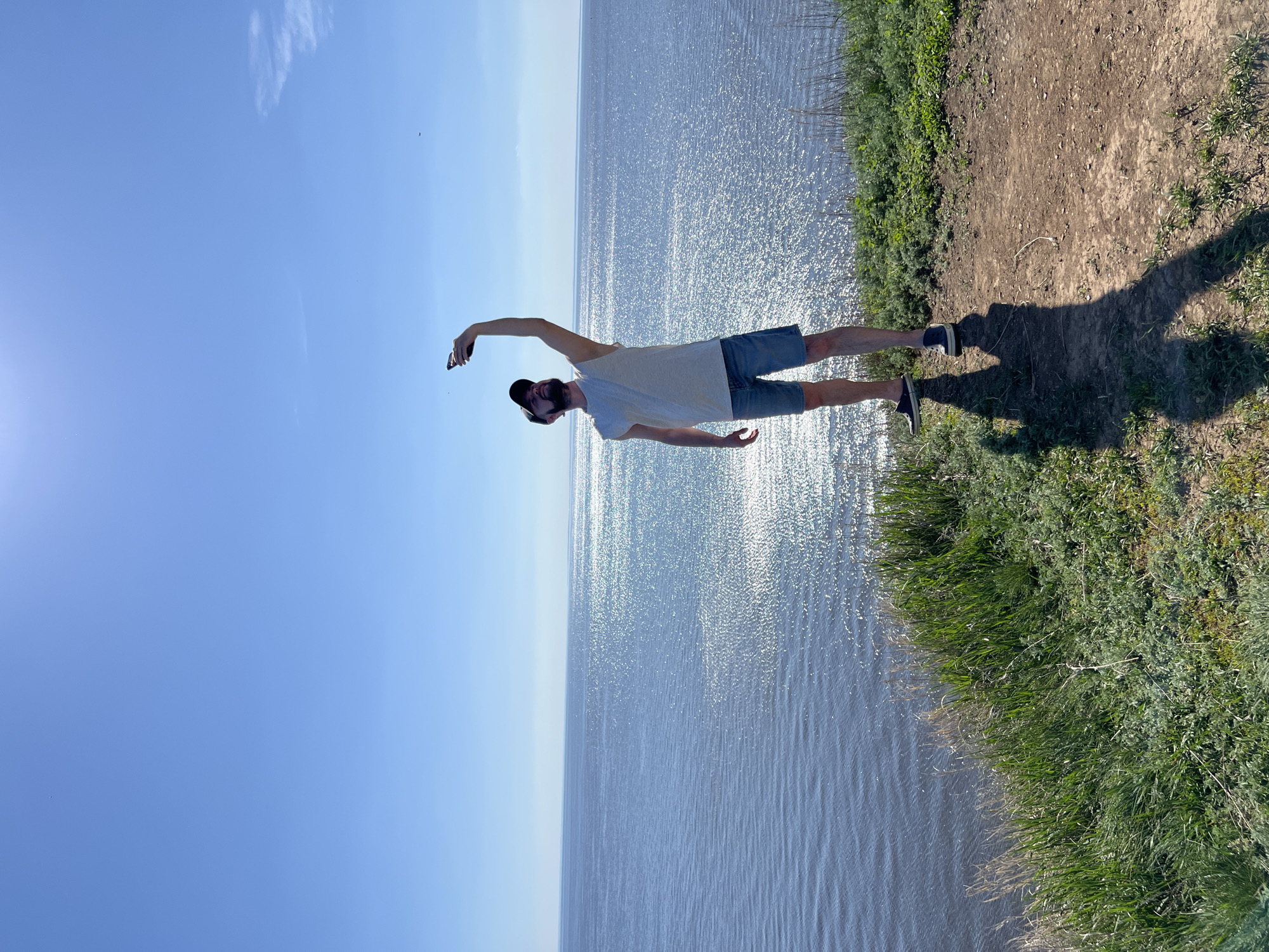 A full body shot of a person on the shore. The person is wearing a white t-shirt, blue shorts, and a black cap. The person has one arm raised, and the other arm is at their side. The water is sparkling in the sunlight. The sky is blue and sunny. The photo was taken outside near Шуран, Russia.