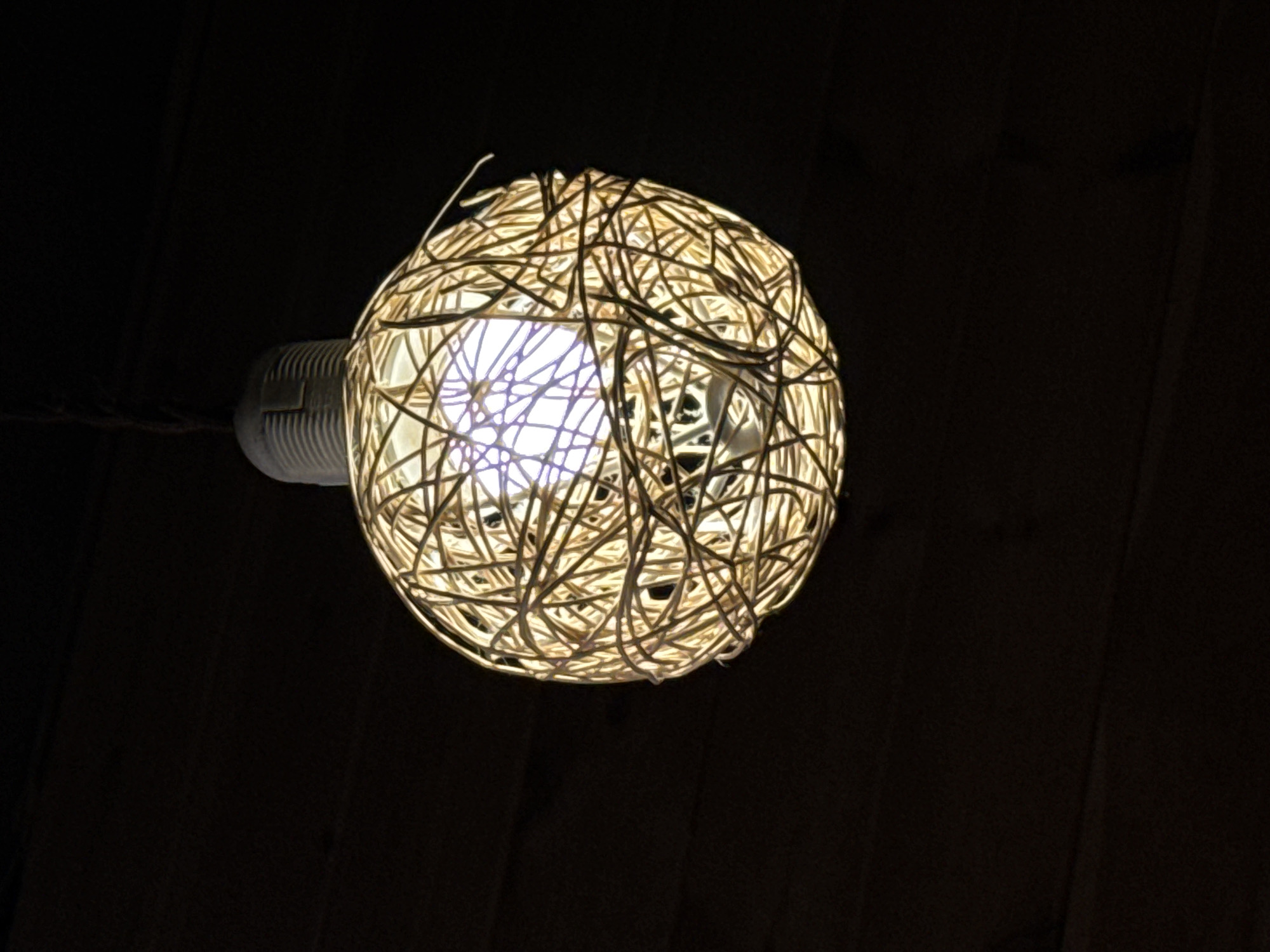 A close-up shot shows an illuminated lightbulb encased in a woven ball of thin, light-colored string or twine. The lightbulb and its holder are visible to the left. The background is dark, appearing to be a wooden surface. The image has a high contrast, with the lightbulb and surrounding woven material strongly highlighted against the darkness. The location is not determinable from the image.