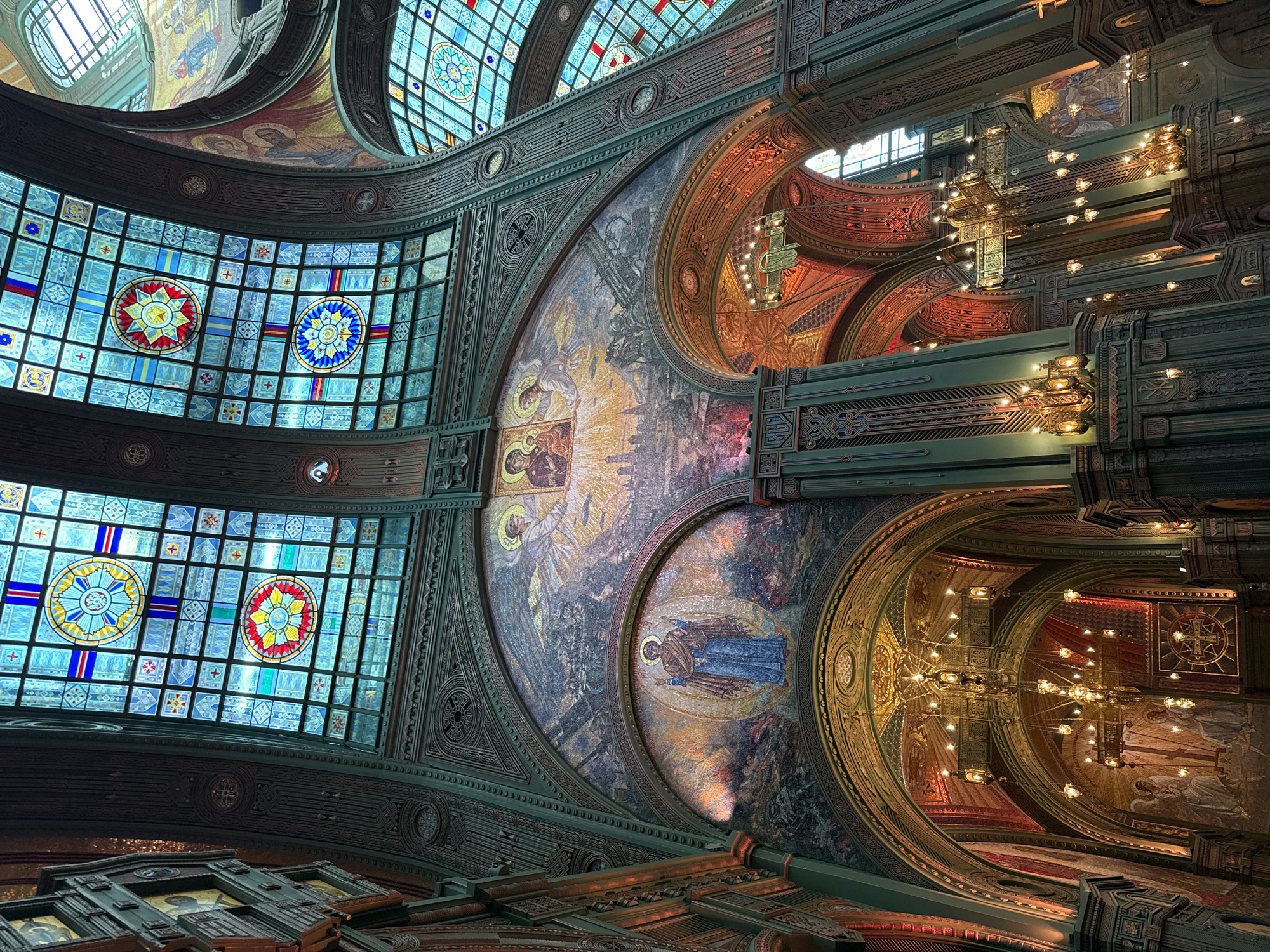 The image depicts the interior of a grand cathedral, characterized by intricate mosaics and vibrant stained-glass windows. The architecture features soaring arches, ornate columns, and elaborate ceiling details. The stained-glass windows are a prominent element, filled with geometric patterns and circular motifs in various colors, primarily blues, yellows, and reds.
Large mosaic murals adorn the curved walls above the arches. One prominent mosaic depicts figures that appear to be religious icons, surrounded by celestial light and possibly scenes of historical or biblical events. Another mosaic features a solitary figure in blue, set against a colorful, textured background. Smaller mosaics and religious imagery are visible in other areas of the cathedral, including on the upper walls and within arched recesses.
The lighting in the cathedral is a mix of natural light filtering through the stained glass and artificial light from numerous chandeliers and wall-mounted sconces. The chandeliers are elaborate, multi-tiered fixtures with many small bulbs, casting a warm, golden glow. The walls and architectural elements are predominantly painted or decorated in shades of green and dark wood tones, contrasted by the rich colors of the mosaics and stained glass. The overall impression is one of opulence, spiritual grandeur, and meticulous craftsmanship.
The cathedral is located in the city of Zelenaya Roshcha, Russia.