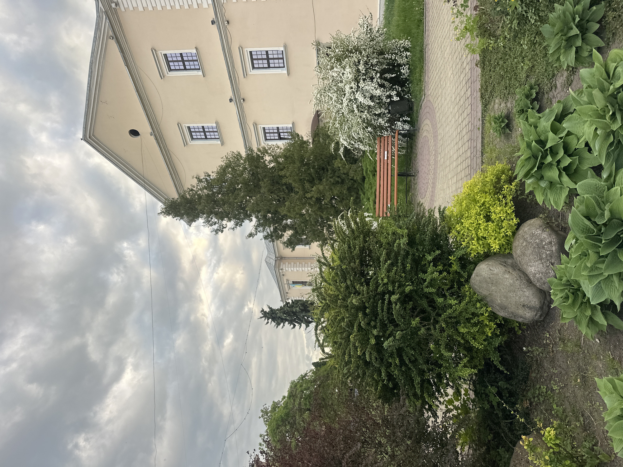 The image depicts a partially visible, light yellow building with three visible windows on its side facade. A bench is situated in front of the building, surrounded by a mixture of shrubs and bushes. A paved walkway runs adjacent to the bench. In the foreground, there are rocks and a variety of green plants. Above, the sky is overcast with heavy clouds. The scene is located in the city of Ternopil, Ukraine.