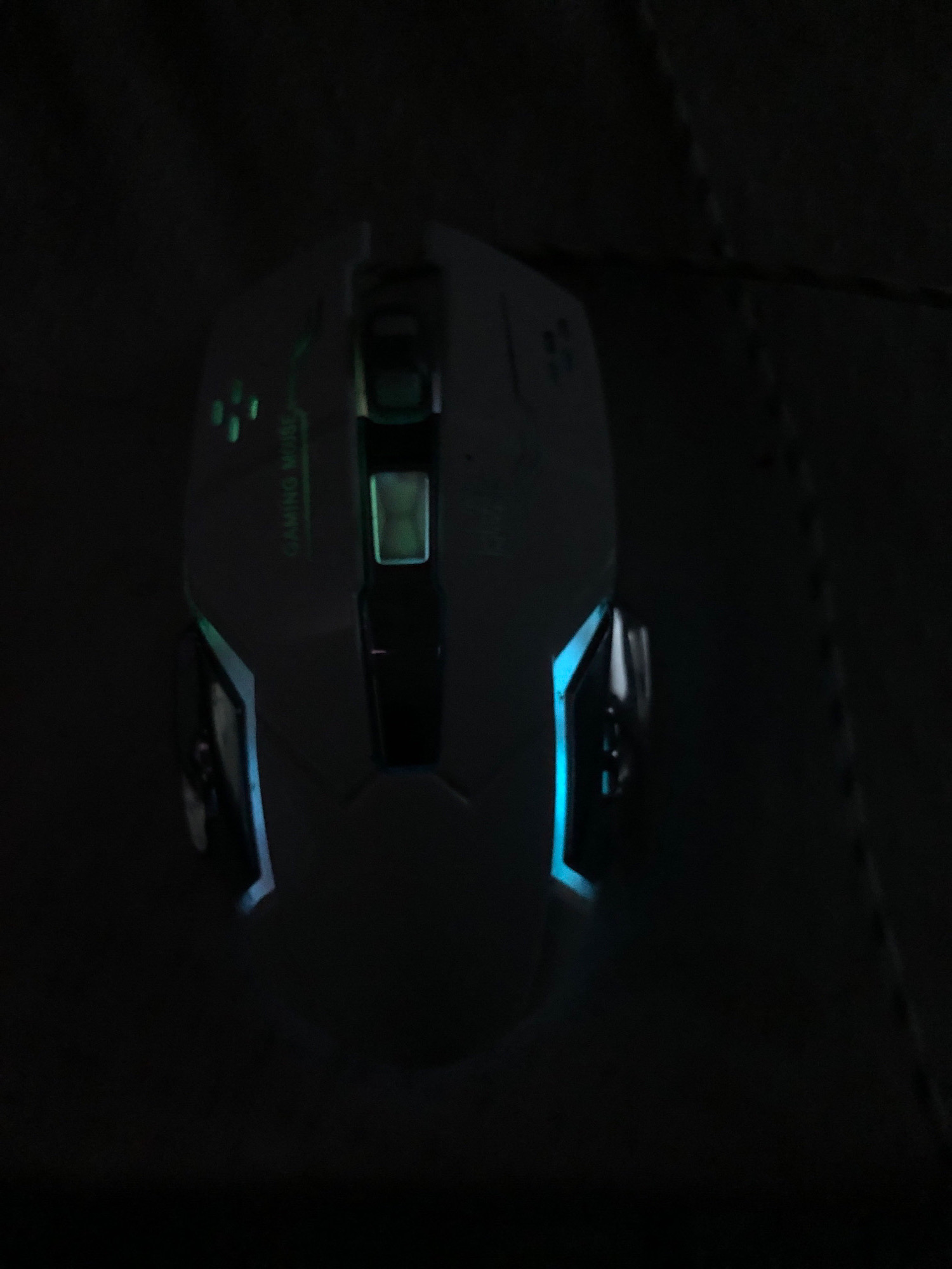A top-down, dimly lit view of a white gaming mouse is visible. The mouse has several illuminated elements, including green lights on the top surface and blue lights along the sides. The words "GAMING MOUSE" are faintly visible in green text on the side of the mouse. The background is dark, with some textured material visible to the right. There are no people or specific geographical landmarks identifiable that would indicate a location in Saidan, Pakistan.