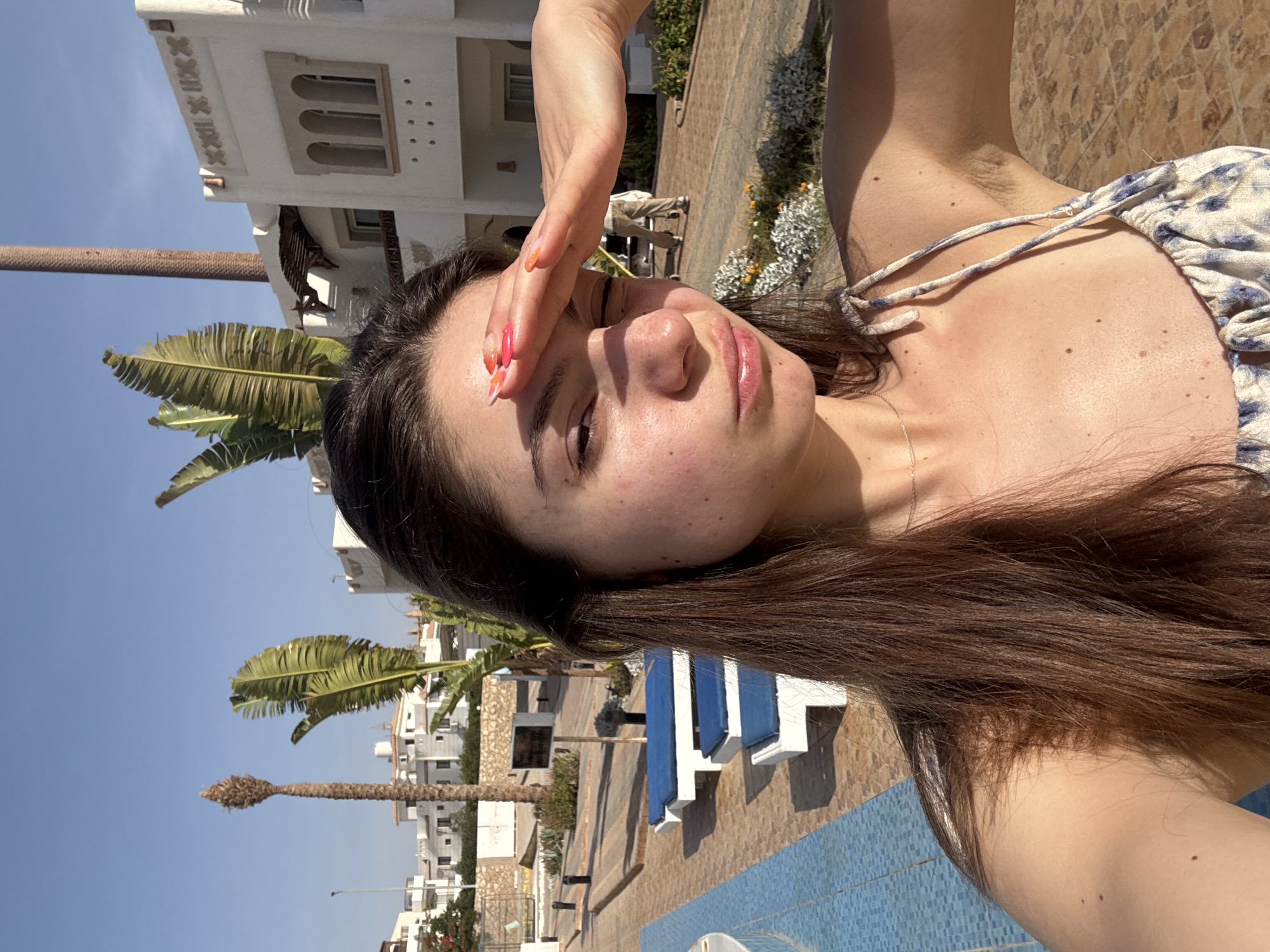 A young woman is posing for a selfie in an outdoor location. She is shielding her eyes from the sun with her right hand. The background shows a resort-style setting with palm trees, buildings, and what appears to be a swimming pool. The city is unknown.