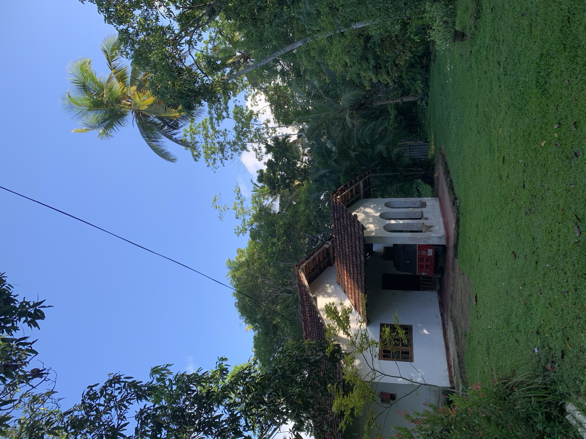 The image depicts a building in Kabalana, Sri Lanka, situated on a plot of land with a grassy lawn. The structure has a white exterior, a red-tiled roof, and windows with wooden frames. The scene is surrounded by lush vegetation, including trees and tropical foliage. A palm tree is visible against a blue sky. A utility line runs across the sky. The image appears to be taken during daylight hours. A red vehicle is partially visible inside the building.