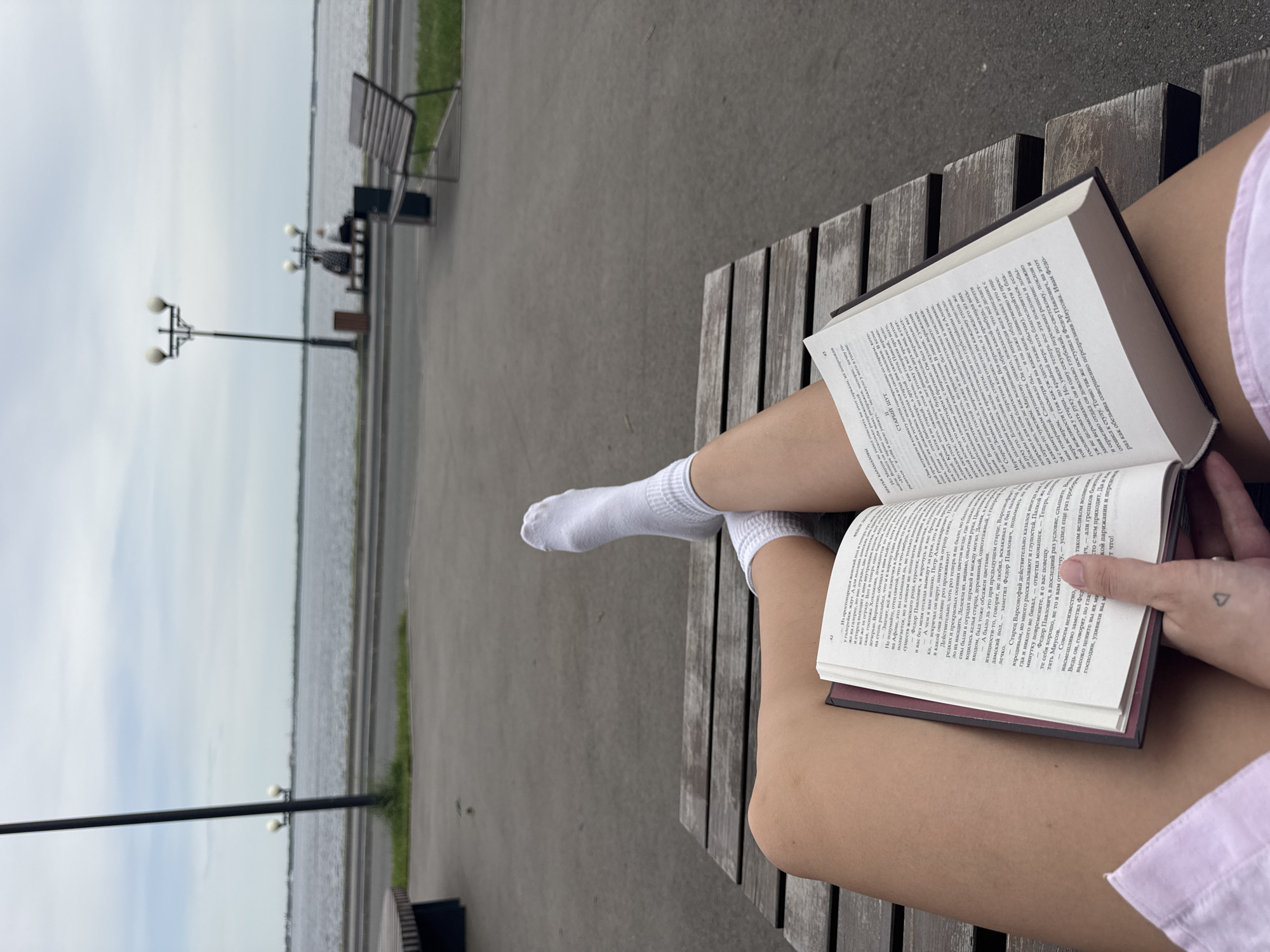 A person is sitting on a wooden bench outdoors, reading a book. The person is wearing white socks and a light pink shirt, and their legs are crossed. The book is open, and the text is in Russian. In the background, there is a gray paved area, streetlights, and a body of water. In the distance, benches line the edge of the paved area. This scene is likely taken in Saratov, Russia.