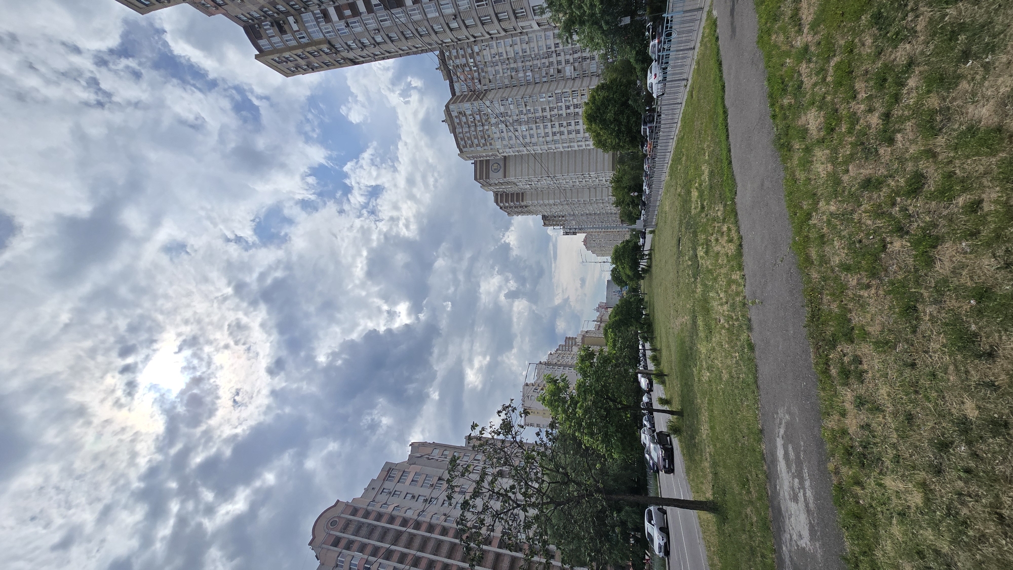 This image depicts a wide urban street, specifically Academician Palladin Avenue in the Sviatoshynskyi District of Kyiv, Ukraine. The scene is viewed longitudinally down the avenue under an overcast sky that exhibits patches of brighter light, suggesting a partially obscured sun, along with areas of visible blue sky.
The avenue is flanked on both sides by rows of tall, multi-story residential apartment buildings that recede into the distance. The buildings on the left side are characterized by a mix of light-colored facades and reddish-brown elements, featuring numerous windows and balconies. The buildings on the right side are primarily light beige and brown, with similar window and balcony configurations; one prominent building on this side displays a distinct circular emblem or decorative motif near its upper section.
A wide, grassy median strip runs down the center of the avenue, separating the two directions of vehicular traffic. This median contains a paved pedestrian pathway and several young deciduous trees with green foliage. Both sides of the avenue are active with cars, some in motion and others parked, comprising various models and colors. The immediate foreground shows a patch of mixed green and dried grass and a segment of a paved path. No people are visible or identifiable within the frame.