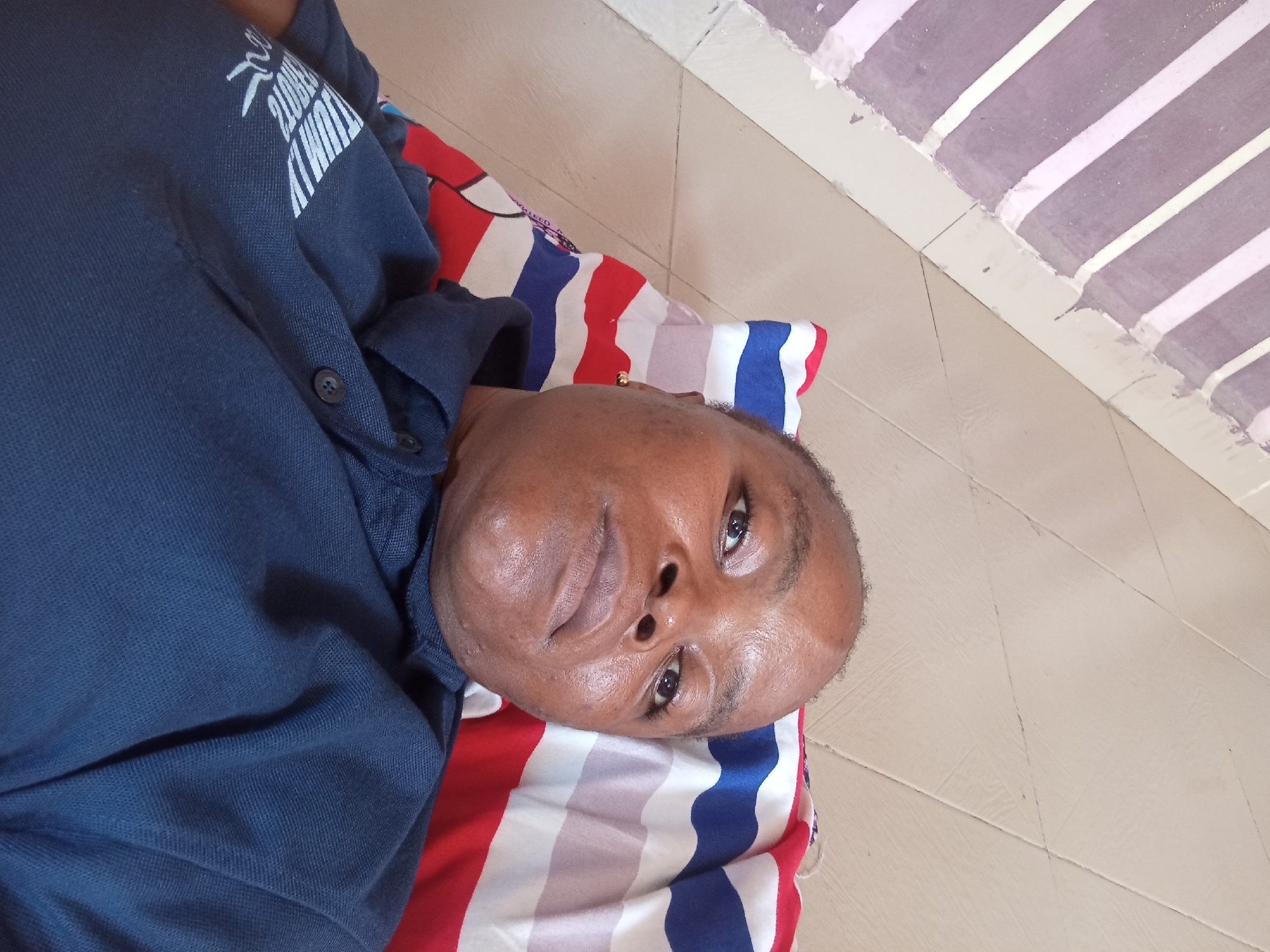 The image shows a person lying down on a striped pillow. The person is wearing a dark blue shirt with white writing on the left sleeve. They have dark skin and short hair. The person is wearing a small gold earring in their left ear. The pillow has stripes of red, white, and blue. The background appears to be a tiled floor with a pattern of light-colored tiles. The lighting suggests an indoor environment. The image is oriented with the person's head to the right.