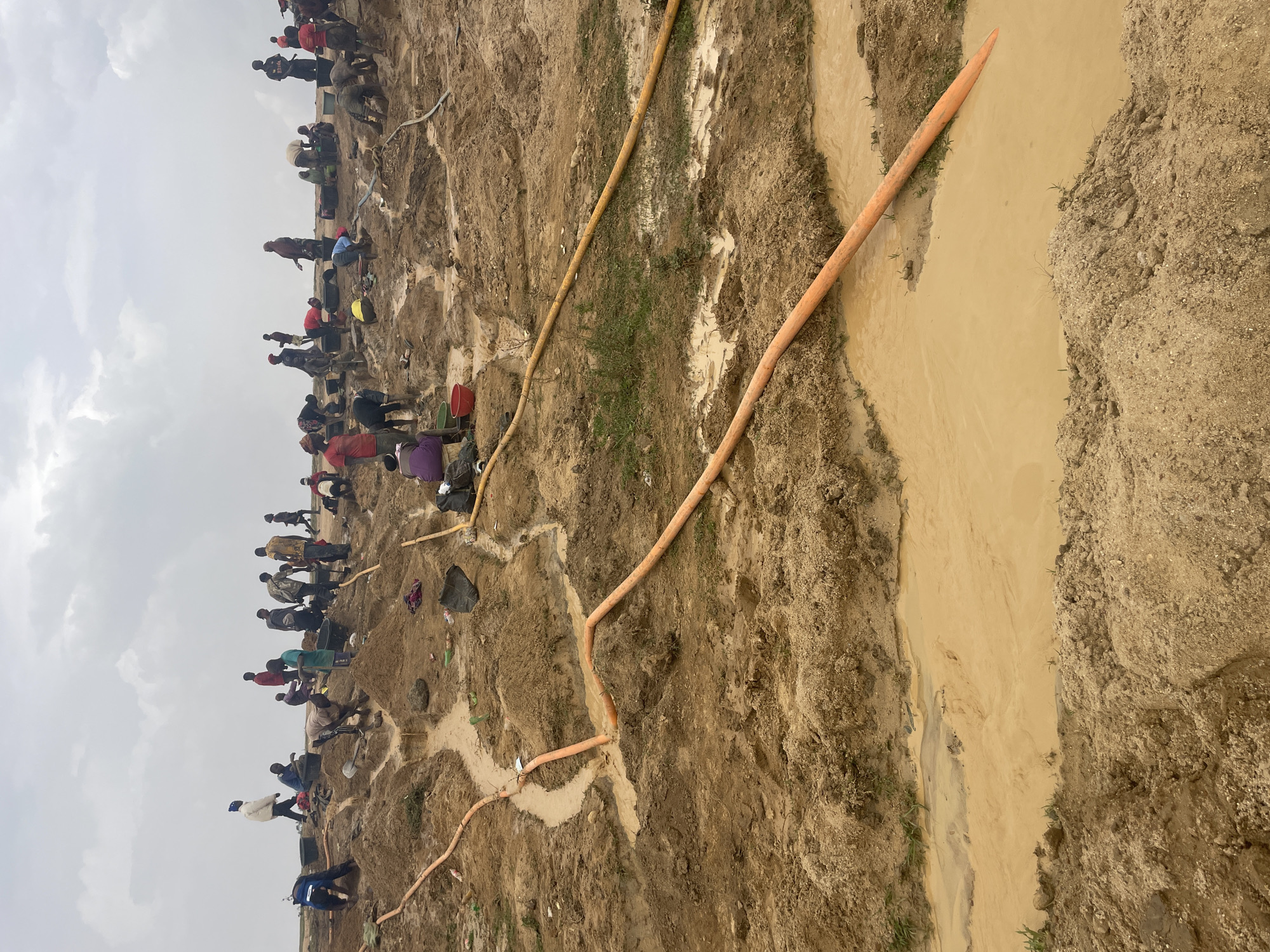 The image depicts an outdoor scene in Maiyanga, Nigeria, showing an area of dirt and mud where a large group of people is gathered. The individuals appear to be working or digging in the dirt. Several orange hoses are visible across the terrain, one leading to a narrow channel of muddy water. The sky is overcast.
