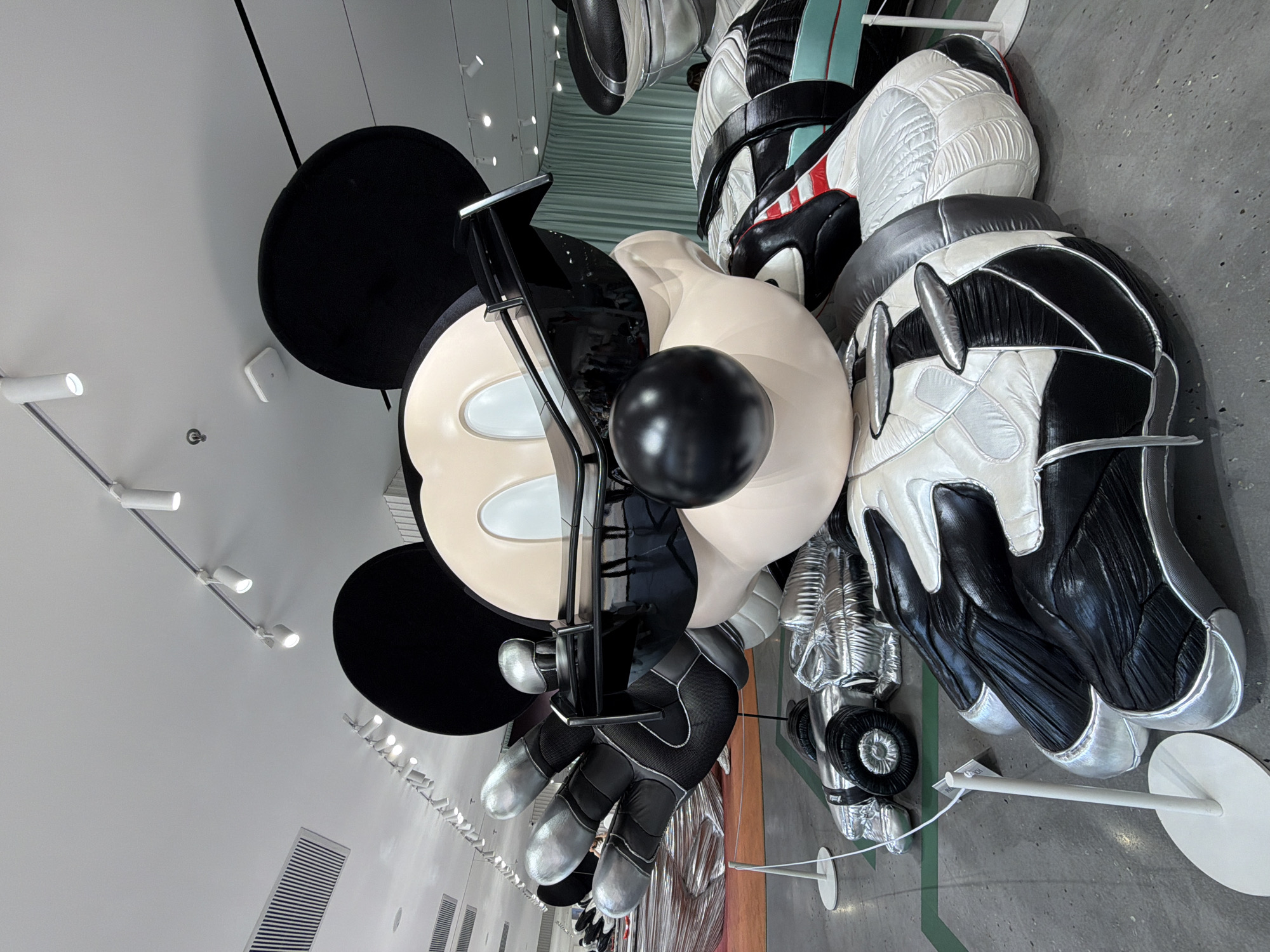 The image depicts a large, three-dimensional art installation of Mickey Mouse in Jing'an District, China. Mickey Mouse is portrayed in a stylized, futuristic manner. The installation is composed of various materials, including black, white, silver, and beige elements. Mickey's head is large and cartoonish, with the iconic ears, face, and a black nose. His gloves are large and metallic-looking, possibly representing robotic or futuristic elements. The rest of Mickey's body is abstracted into a series of segmented, balloon-like forms, some with racing suit details. The sculpture is displayed indoors on a gray concrete floor in front of a sheer curtain. Lighting fixtures are visible on the ceiling.