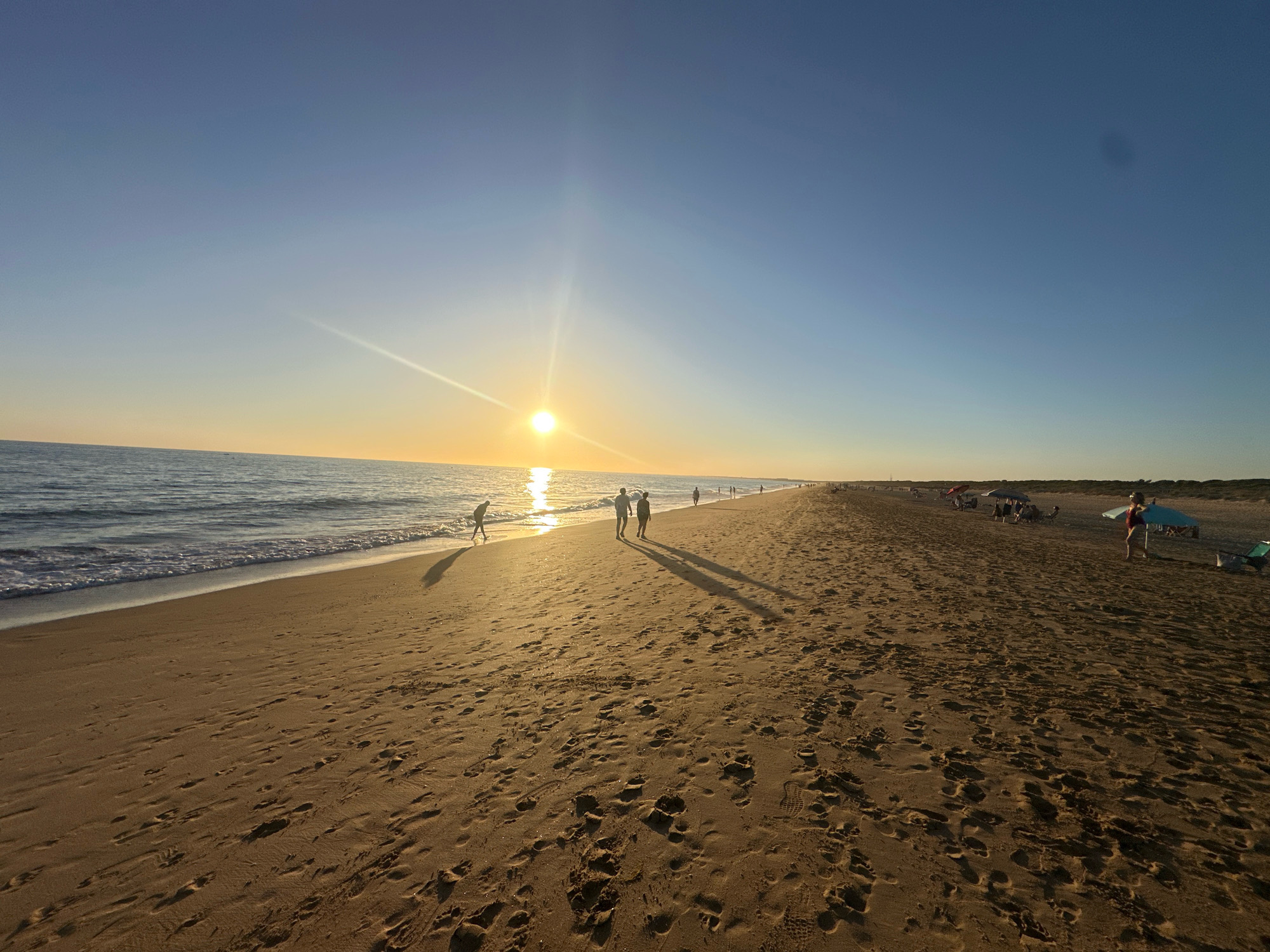 The image depicts a wide, sandy beach with footprints scattered across the surface. The ocean is to the left, with gentle waves lapping the shore. The sun is low in the sky, casting a warm, golden light and creating a bright glare. A few individuals are visible walking along the beach, some near the water's edge and others further up on the sand. In the distance, more people and beach umbrellas suggest a populated area. The sky is a clear gradient of blue, transitioning to a lighter hue near the horizon where the sun is setting. The scene appears to be the beach at Punta Umbría, Spain, known for its extensive sandy shores and popular tourist season.