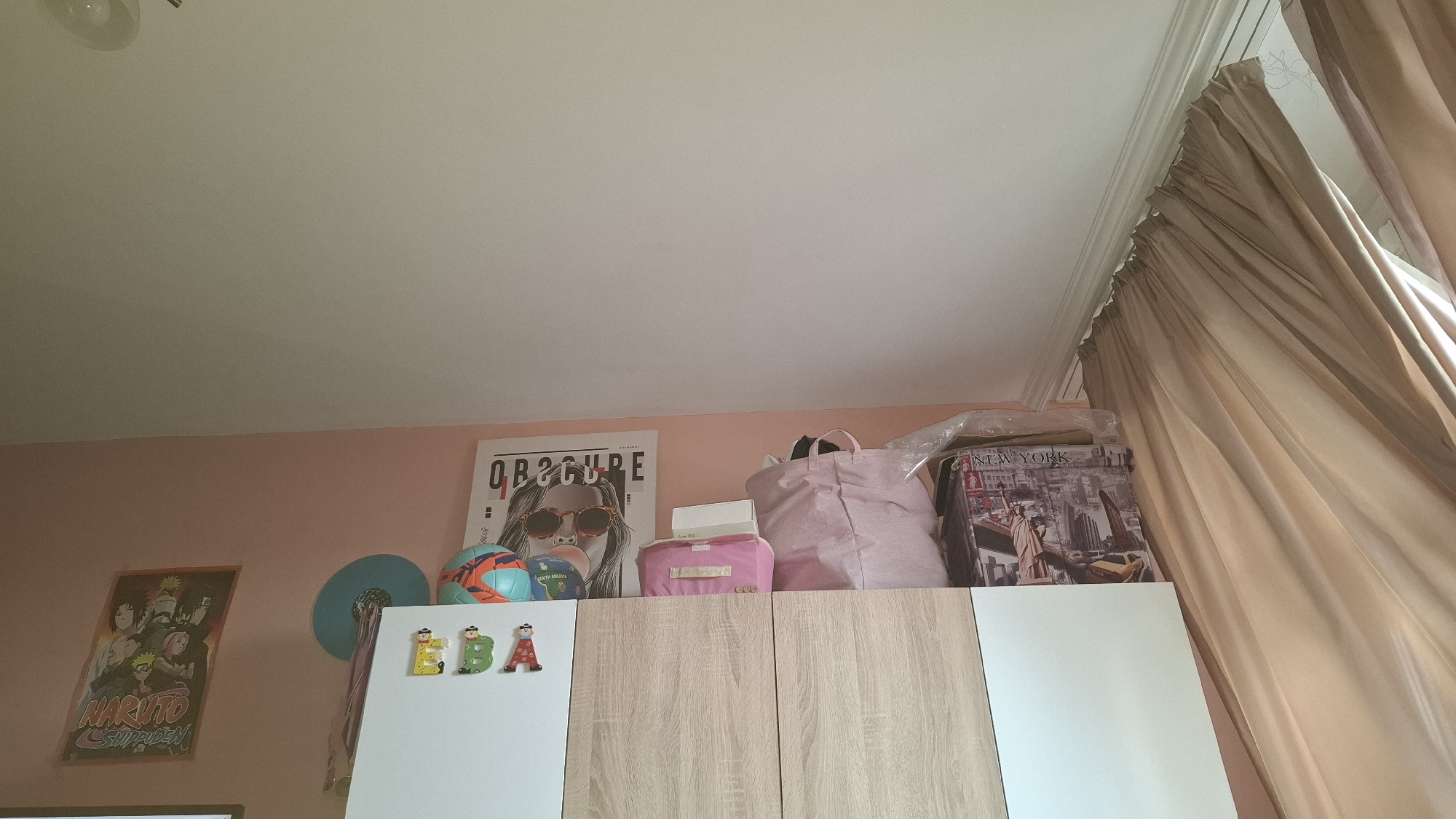 The image depicts the interior of a room, likely a bedroom. The walls are painted a light pink color. A wardrobe with a combination of white and light wood grain doors is in the foreground. The doors are adorned with fridge magnets spelling out "EBA". On top of the wardrobe are various items, including a soccer ball, a pink tote bag, a small pink box, a poster featuring a person with sunglasses and the word "OBSCURE", and a puzzle box depicting New York City. On the left wall, there is a poster featuring characters from the anime Naruto. The curtains are heavy and beige, partially obscuring a window. The location is Sofia, Bulgaria. There are no people visible in the image. The objects are arranged on top of the wardrobe, suggesting storage.