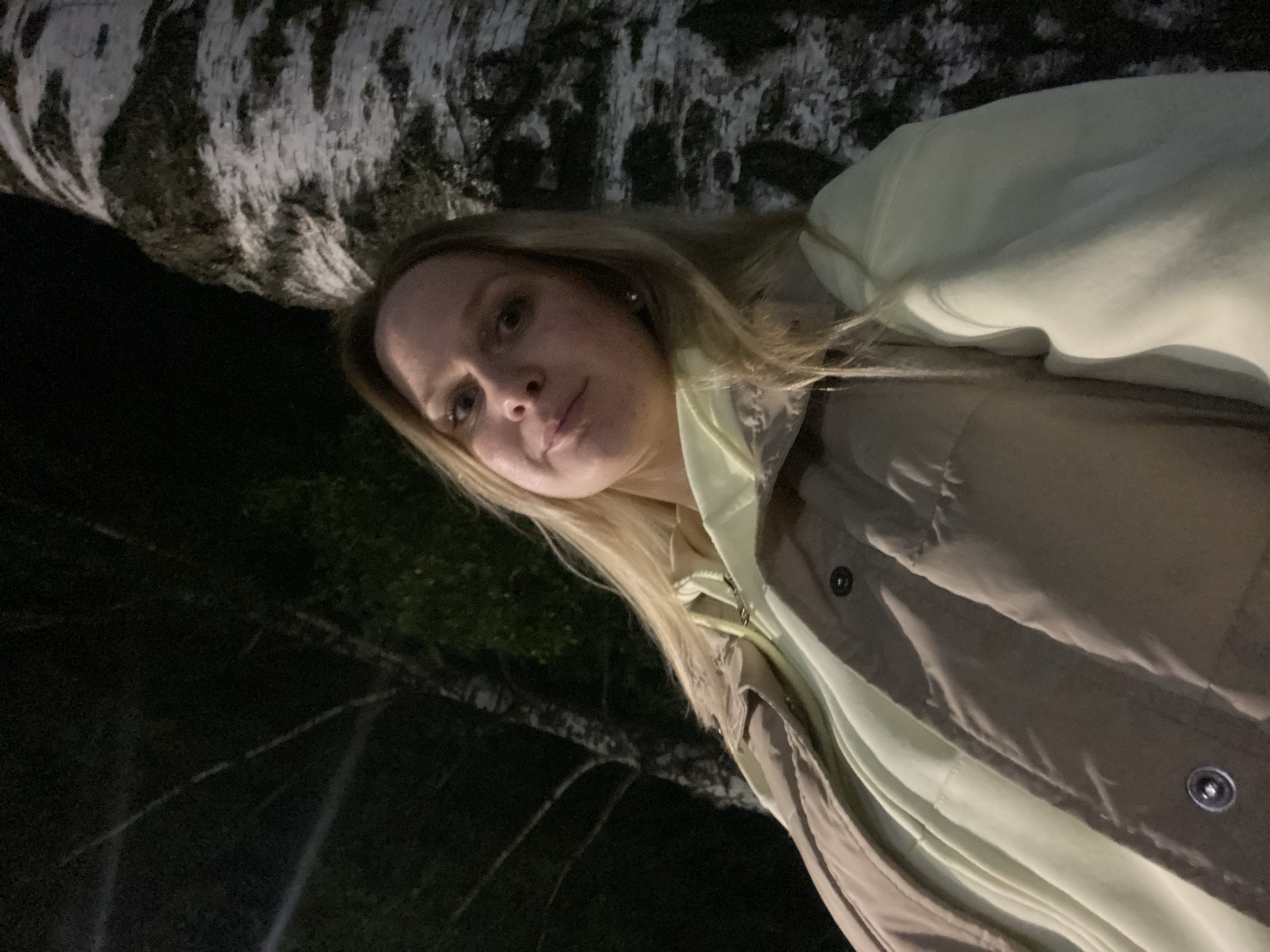 An eye-level, outdoor, medium shot features a young woman with blonde hair and fair skin. She is positioned to the left of a large, textured tree trunk that dominates the right side of the frame. She is wearing a pale yellow hooded sweatshirt and a beige puffer vest. The background is dark, suggesting nighttime, with the faint outlines of trees and foliage visible. The overall lighting is dim, with some light source illuminating the woman and the tree trunk. The location is identified as Chaikovsky, Russia.