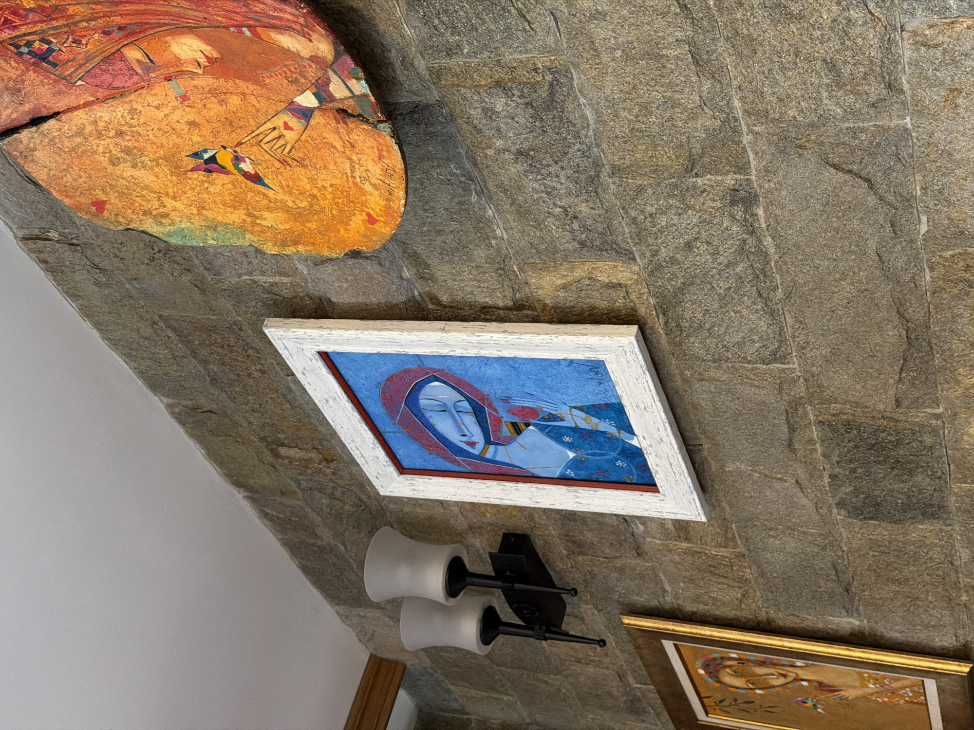 The image shows a stone wall in Кирково, Bulgaria, with several framed artworks hanging on it. On the upper left, there is a large, irregularly shaped artwork in shades of orange and brown. Below it, a rectangular painting with a white frame depicts a woman in a blue and red headscarf. Below this painting, there is a wall sconce with two white cylindrical lights mounted on a black bracket. To the lower right, a portion of another framed artwork is visible, featuring golden hues. The scene is indoors, possibly in a home or gallery.