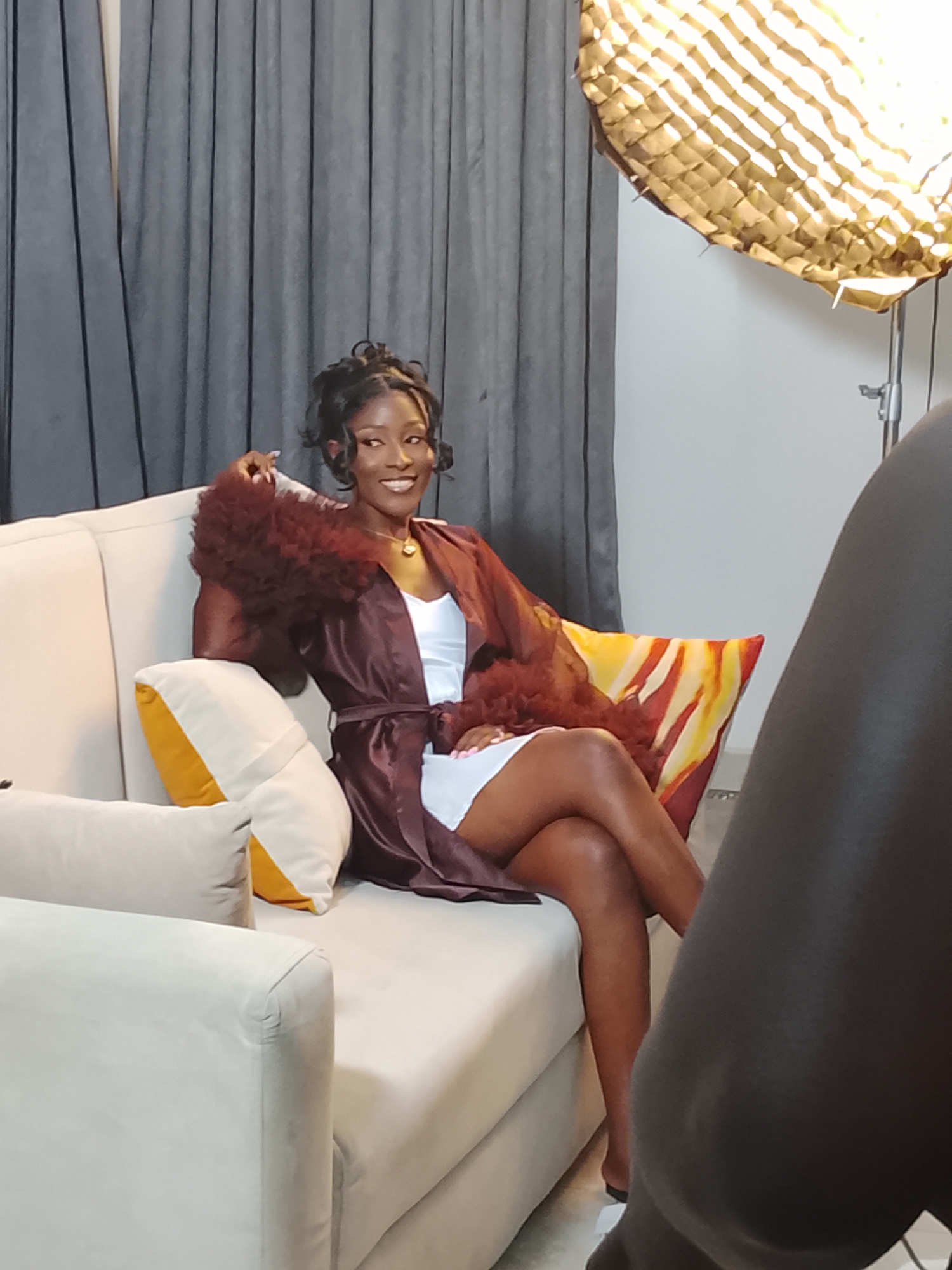 A woman is seated on a light-colored couch. She is wearing a white dress and a dark, fluffy robe, crossed at the knees, smiling and posing for a photograph. The couch has yellow and orange decorative pillows. A gray curtain hangs behind her and off to the right is a large, covered lamp on a stand. In the foreground on the right side of the image, there is a large out-of-focus form of a dark-skinned person, potentially a photographer. The photo appears to be taken indoors, possibly in a studio in Gwarinpa, Nigeria.