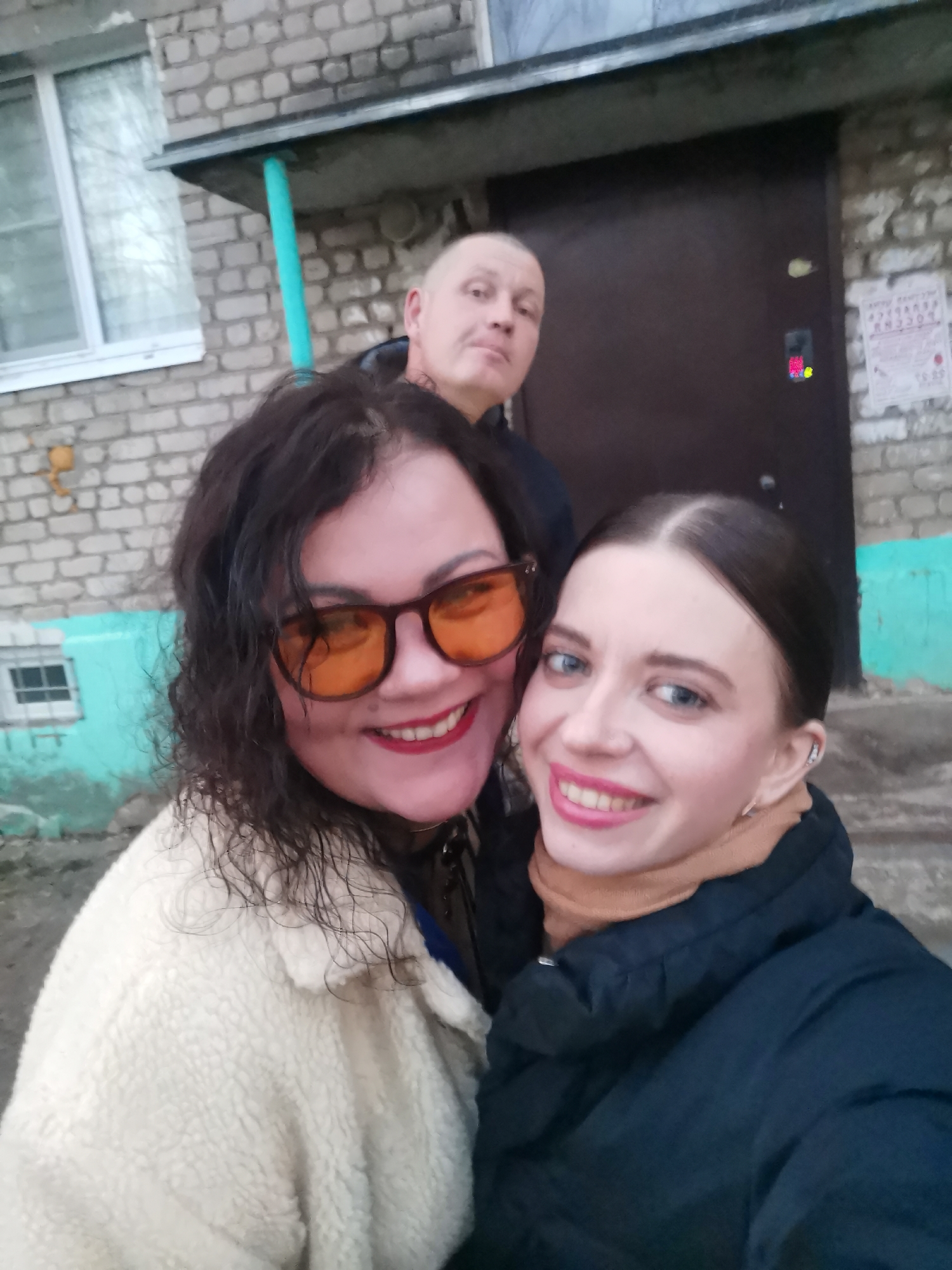 A group of three individuals, two women in the foreground and a man in the background, are captured in a photo outdoors, likely a selfie. The two women are positioned closely together, both smiling broadly at the camera. The woman on the left has dark, curly hair and wears large, distinctive orange-tinted sunglasses, bright red lipstick, and a cream-colored, textured fluffy jacket. The woman on the right has dark hair pulled back, bright blue eyes, pink lipstick, and is dressed in a black puffer jacket over a tan turtleneck or scarf. In the background, partially obscured, a man with very short or bald hair looks towards the camera with a subtle expression, wearing a dark jacket.
The setting is in front of a weathered brick building, possibly an apartment complex or a house. A white-framed window is visible on the left side of the brick wall. In the center-right, there is a dark brown entrance door equipped with an electronic intercom or keypad, which displays some illuminated red and yellow digits or symbols. To the right of the door, a piece of weathered paper with illegible Cyrillic text and the number "10.00" is affixed to the brickwork. A section of the building's exterior, including a pipe, is painted a distinctive turquoise blue. The lighting is soft and diffused, suggesting an overcast day, and the warm clothing worn by the individuals indicates cool or cold weather. The overall mood conveyed by the women's smiles is cheerful.