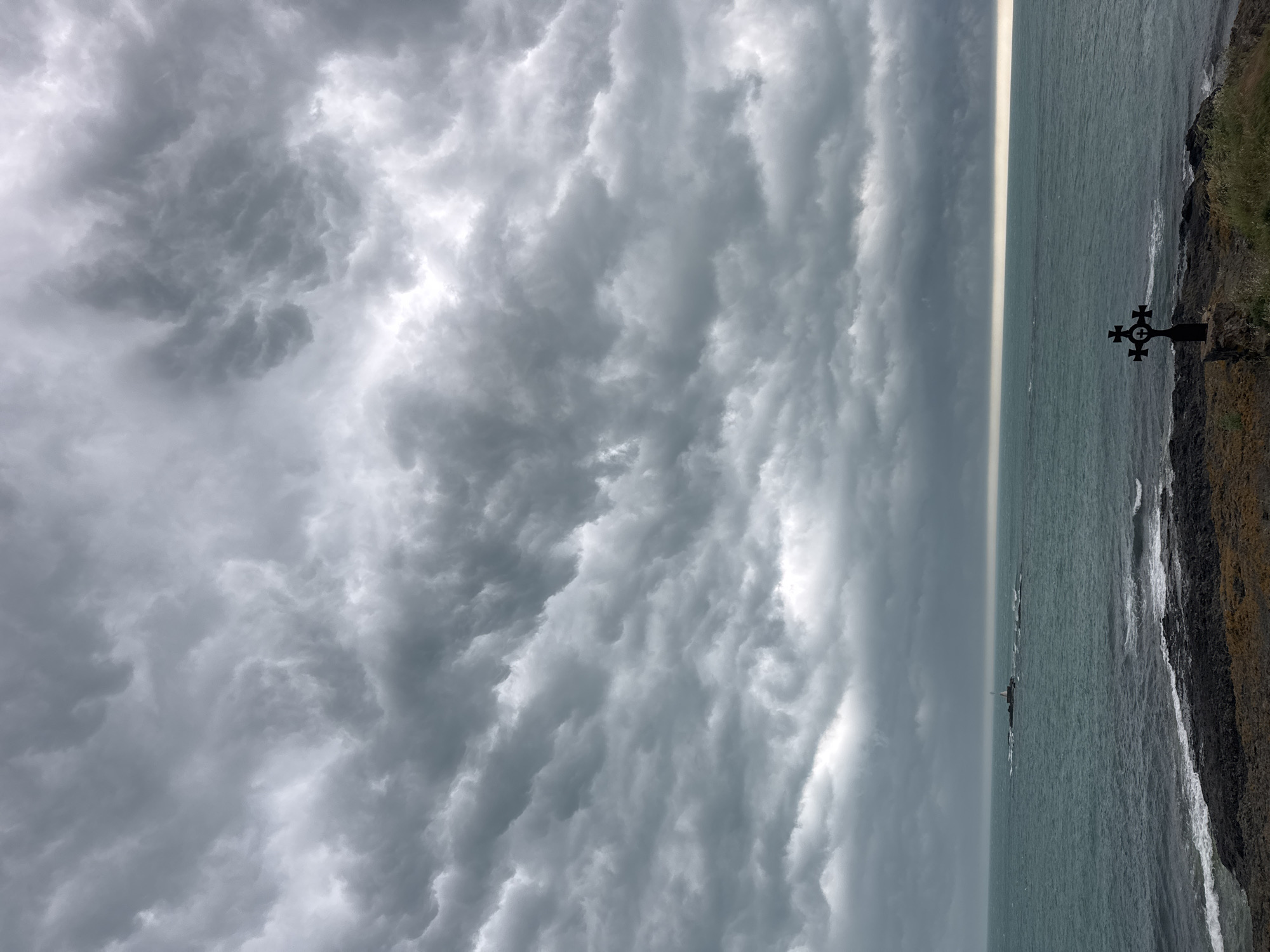 The image, rotated on its side, depicts a dramatic coastal landscape under a heavily clouded sky. When viewed upright, the upper two-thirds of the frame are dominated by an expanse of turbulent, dark grey clouds, exhibiting intricate textures and deep shadows, indicative of stormy or impending stormy weather.
Below the tumultuous sky, a wide body of dark, grey-green sea stretches towards the horizon. The water appears choppy, with visible waves breaking near a rugged, dark rocky coastline that occupies the right side of the frame. A narrow, bright strip of light is visible along the horizon line where the sky meets the sea, possibly a distant break in the clouds or a reflection.
On the rocky coastline in the lower right, a small, ornate black metal cross with circular elements is prominently featured. Further out in the sea, a small white structure, likely a lighthouse or navigational beacon, stands on a rocky islet. The land itself is dark and rocky, with sparse patches of dry-looking vegetation.
No people or specific activities are visible. The scene conveys a somber, dramatic, and wild atmosphere, characteristic of an overcast day on a rugged coast. No visible text is present. The setting is consistent with a coastal area like Tsarevo, Bulgaria.