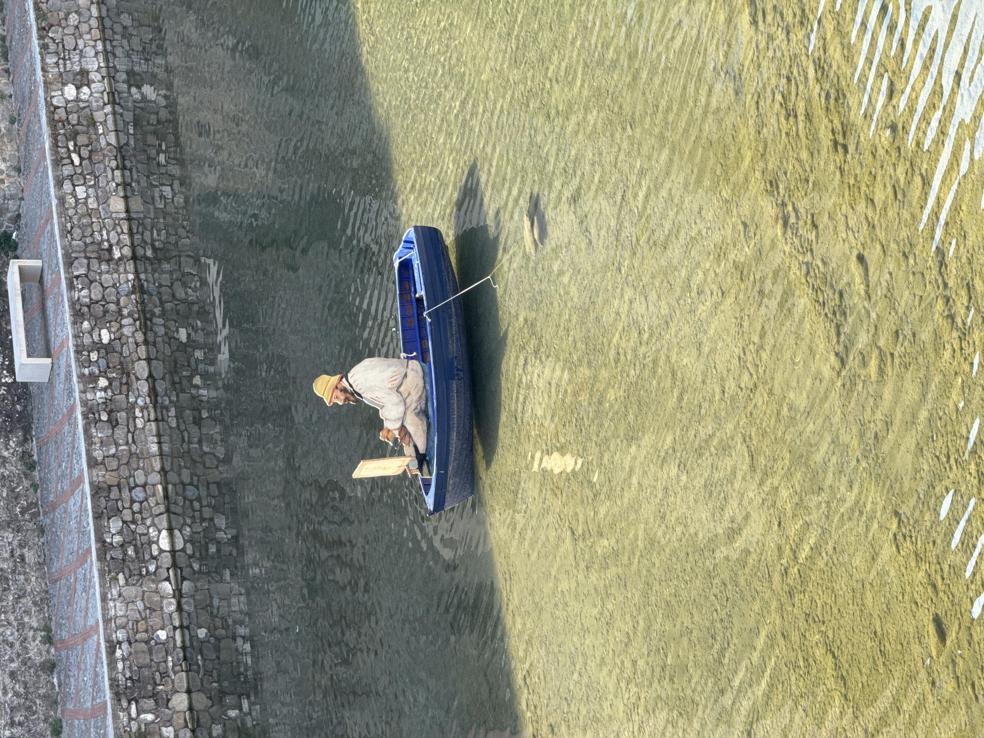 In Dolceacqua, Italy, a man wearing a straw hat and a light-colored jacket is seated in a blue rowboat in the shallow, clear water of a river. He appears to be painting on a canvas or easel positioned in front of him. The riverbed is visible beneath the water, with some rocks and aquatic vegetation. The river is bordered by a stone wall and a paved walkway, with a white bench visible on the walkway in the background. The scene is illuminated by natural light, casting shadows on the water.