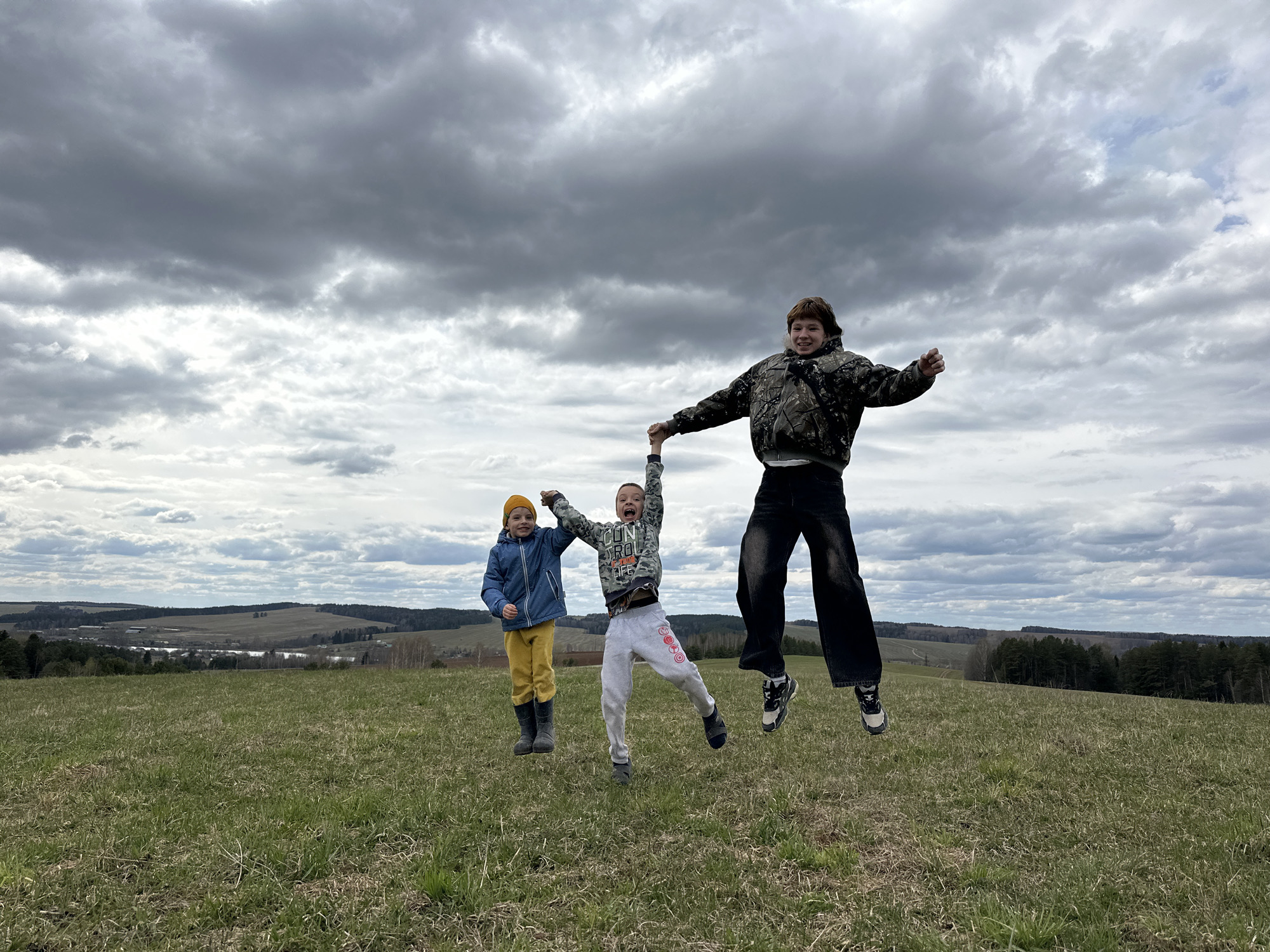 Three children are jumping in a grassy field under a cloudy sky. The child on the left wears a yellow beanie, a blue jacket, and yellow pants with black boots. The child in the center is wearing a gray sweatshirt, gray sweatpants, and black boots. The child on the right is wearing a camo jacket, a white undershirt, black pants, and two-toned sneakers. The children are holding hands as they jump in unison. The scene appears to be in a rural area, possibly near the city of Альняш, Russia, based on the surrounding landscape of rolling hills and forests in the background.