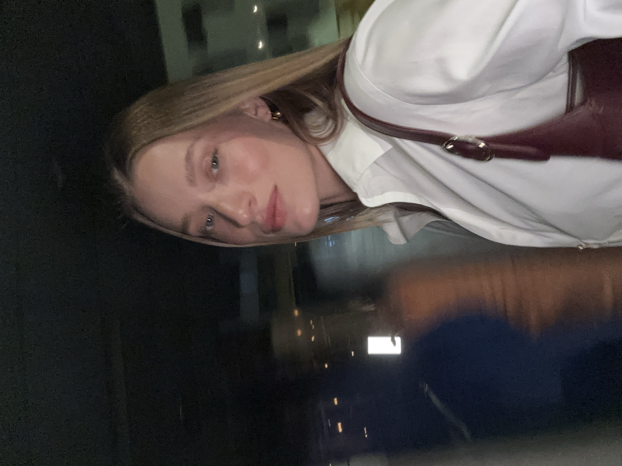 The media file shows a close-up of a young woman with blonde hair, tilted slightly sideways, looking directly at the camera. She is wearing a white button-down shirt and a burgundy harness or vest. Her expression appears neutral, with her makeup subtly highlighting her features. Her hair is long and straight, falling around her face.
The background is dark and appears to be at night, with some blurry lights suggesting an urban environment. There are reflections in what might be a window or a polished surface in the foreground, showing a blurred cityscape with lights and some indistinct shapes. The overall mood is intimate due to the close-up framing, but the blurry background creates a sense of distance. The lighting is somewhat dim, with the focus on the woman's face. There are no visible signs or text. The presence of artificial lights in the background indicates it is likely nighttime. Given the location context of Tel-Aviv, Israel, this could be a selfie taken in the city at night.
