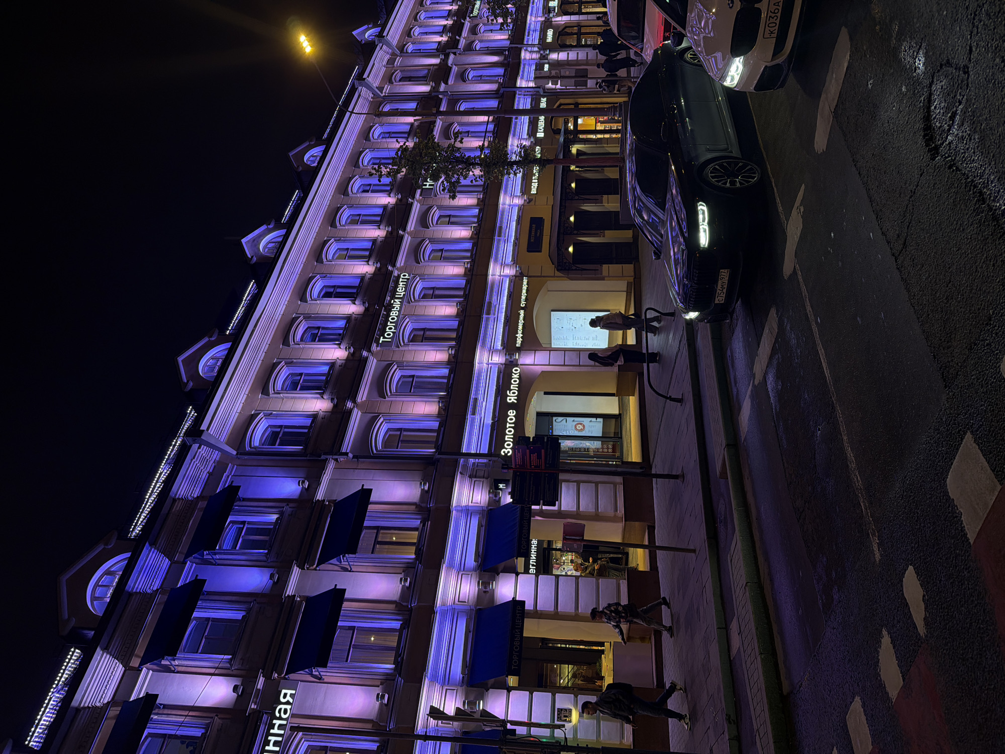 At night, a multi-story building is illuminated with blue and pink lights in Moscow, Russia. The building's facade has multiple windows, awnings, and signs. Signs in Russian include "Торговый центр" (Shopping Center) and "Золотое Яблоко" (Golden Apple), indicating a perfumery. People walk on the sidewalk in front of the building. Cars are parked along the street. Street lamps provide additional lighting. The road has painted white markings and red areas. Car license plates "C754MY 977" and "K036A" are visible.
