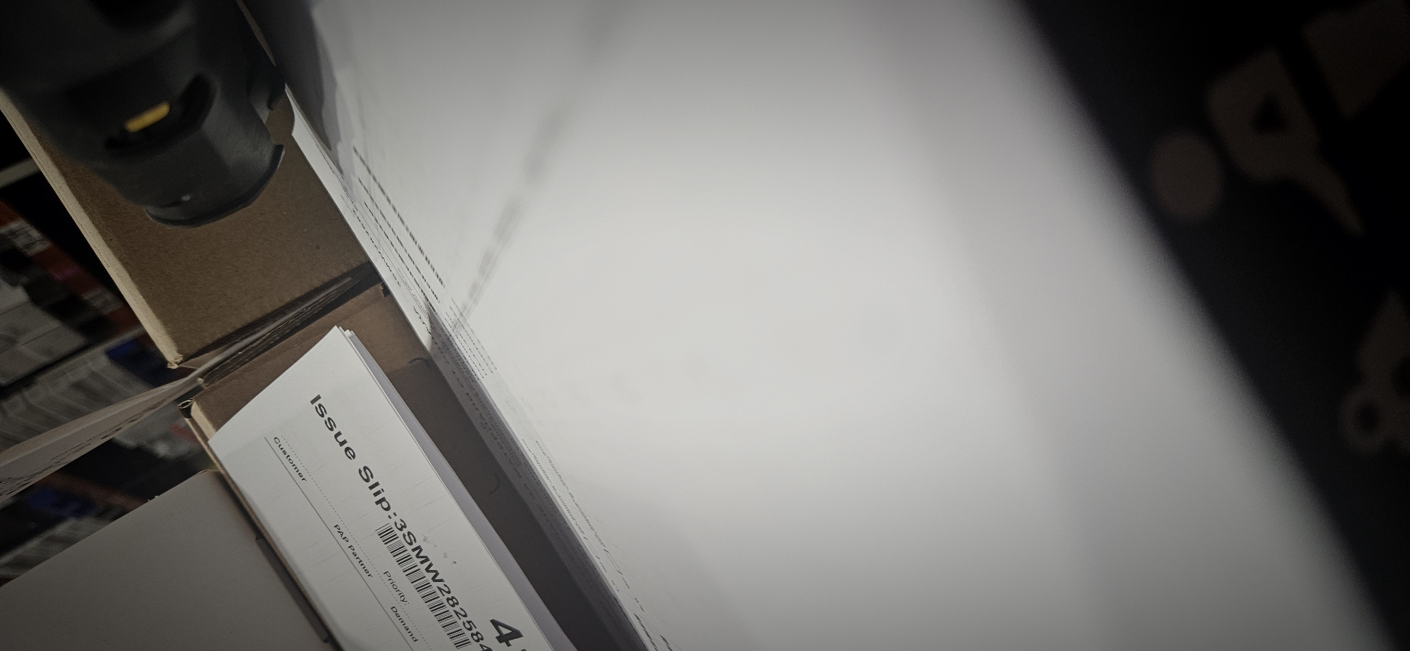 The image, rotated counter-clockwise, captures an indoor scene dominated by documents and packaging materials, likely within a warehouse, office, or commercial setting in Plovdiv, Bulgaria.
In the lower-left quadrant, a stack of white papers features a prominent "Issue Slip." Clearly visible text on this slip includes "Issue Slip:", "Customer", "PAP Partner", "Priority", and "Demand." Below the "Issue Slip:" heading, a barcode is displayed, followed by the alphanumeric sequence "3SMW282584" and the numeral "4".
Resting on a brown cardboard box in the upper-left corner is a black and yellow object, which appears to be a part of a tool or device, possibly a scanner or a component of an assembly line. Other brown cardboard boxes are partially visible around the documents and the black object.
The majority of the right side and upper portion of the image is occupied by a large, brightly lit white surface, likely another box or a workstation, creating a strong contrast with the darker areas. In the far upper-right corner, a dark, blurry background shows faint, indistinct shapes, possibly reflecting obscured text or logos on a dark surface. The lighting is bright and even, characteristic of artificial indoor illumination. No individuals are present in the frame, and the scene suggests an environment focused on inventory, logistics, or administrative tasks.