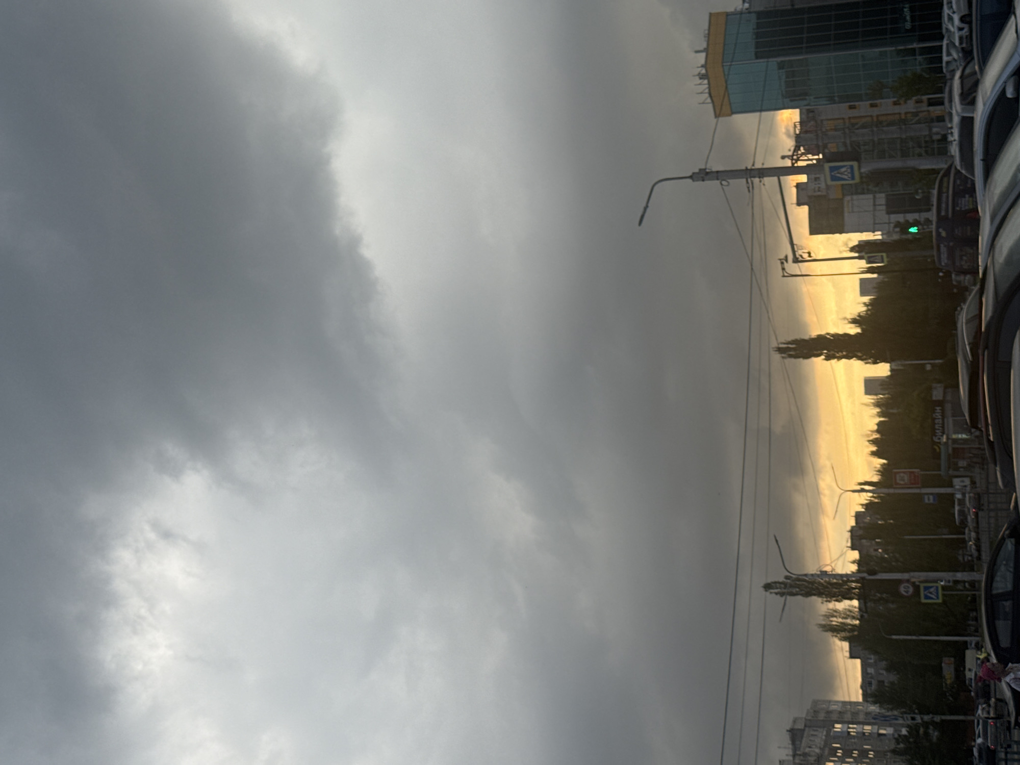 This is a street-level, eye-level, full shot of an urban scene in Ufa, Russia, during what appears to be twilight. The sky is dominated by heavy, dark gray clouds, with a sliver of bright orange and yellow sunset visible near the horizon. This suggests either the sun is setting or has just set.
In the foreground, there is a street with several cars parked and driving. The vehicles are of various types and colors, and the street appears to be congested. There are also several tall, slender trees that are silhouetted against the sky.
In the mid-ground, there are buildings, including multi-story apartment buildings and a modern glass-fronted building with a yellow top. Streetlights and power lines crisscross the scene. There are also pedestrian crossings and street signs visible. One sign has the number "40" on it, likely indicating a speed limit. Another sign displays a pictogram of a person crossing the street.
A person with a pink hat is visible on the sidewalk in the lower left portion of the image, walking towards the left. There is also a kiosk with "Билайн" (Beeline, a Russian mobile operator) signage visible.
The overall atmosphere is somewhat somber due to the heavy clouds, but the vibrant sunset hues offer a contrasting element. The scene depicts a typical urban environment experiencing the transition from day to night under a moody sky.