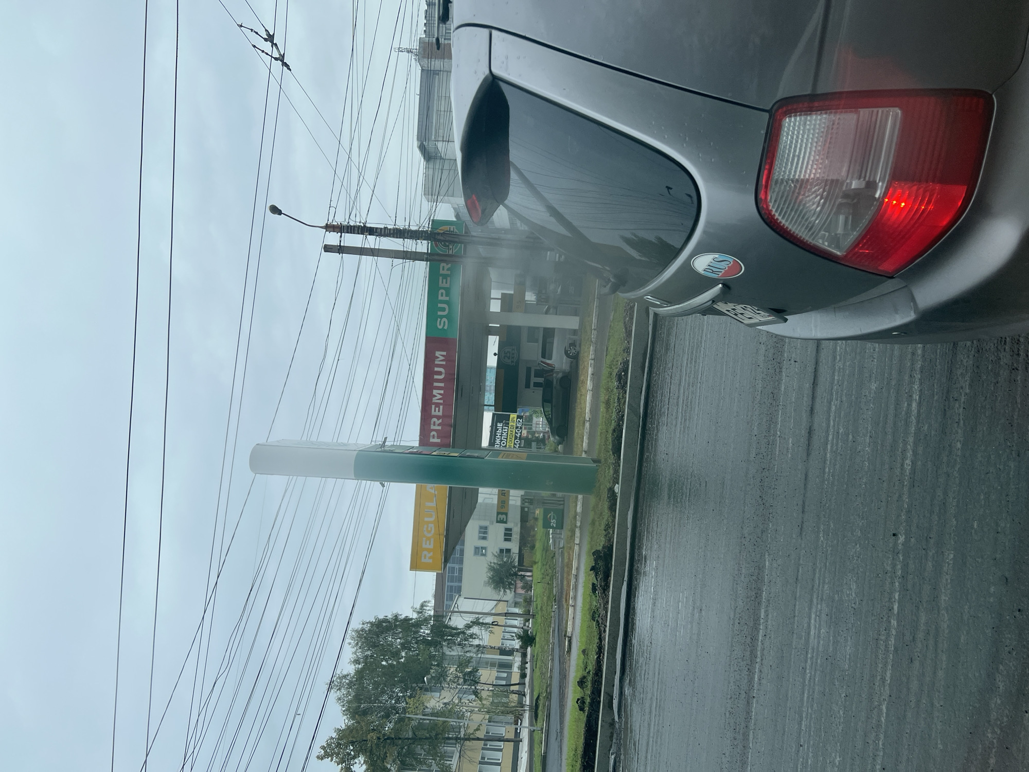 The image depicts a view from inside a car, looking out towards a gas station on a cloudy, overcast day. The car's rear-view mirror and taillight are visible in the foreground, along with a "RUS" sticker on the bumper. The gas station features prominent signage in Russian, indicating different fuel grades like "PREMIUM" and "SUPER," along with pricing information for "98 ДТ" (likely diesel fuel) and a discount promotion for "до 70%" (up to 70%). Overhead, a complex network of electrical wires crisscrosses the sky, typical of urban or suburban environments. The wet asphalt suggests recent rain, and the overall atmosphere is subdued and gray. The scene appears to be a street view in Krasnoyarsk, Russia, based on the provided location context and the "RUS" sticker. There are no people clearly visible, and the focus is on the roadside infrastructure and the car's interior elements.