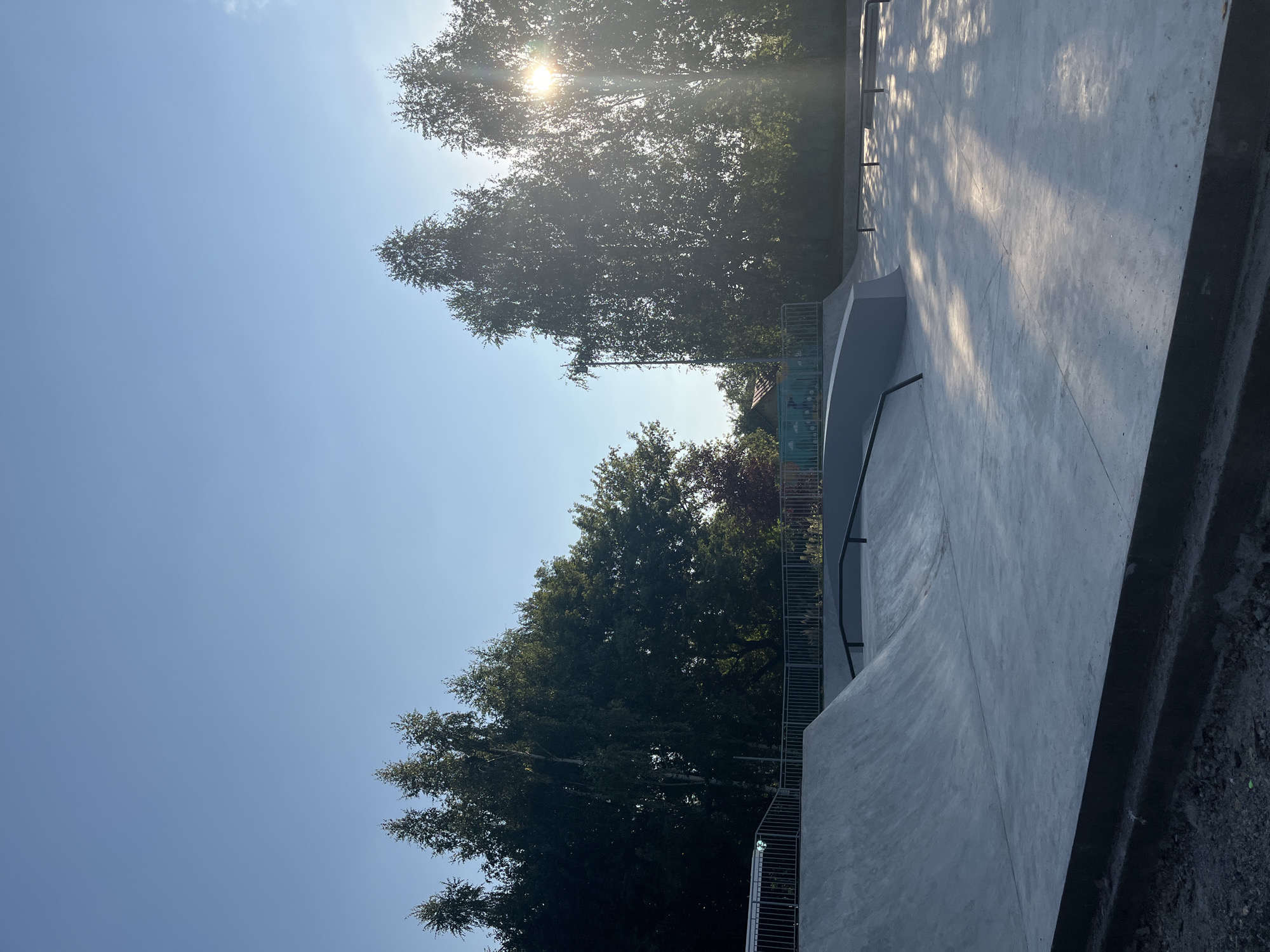 An outdoor concrete skatepark is shown under a clear blue sky, with the sun partially obscured by trees, casting long shadows across the park. The skatepark features various ramps, bowls, and rails, with a metal railing surrounding some of the elevated areas. In the background, a dense line of trees forms a backdrop, with a small building visible to the left of the trees. The ground in the foreground is rough and unpaved, with construction material visible. The overall scene depicts a modern recreational facility in a natural setting.