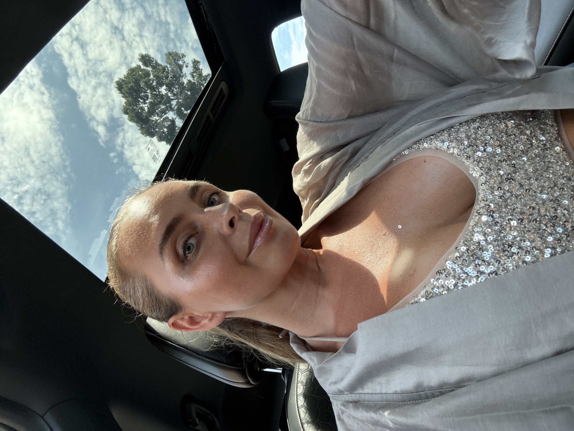 In an indoor vehicle setting, a fair-skinned woman with blonde hair pulled back in a ponytail is seen from the chest up, looking towards the camera. She is wearing a sparkly, silver tank top and an open, light-colored silk robe. Sunlight illuminates her face and chest. The car's sunroof is open, revealing a blue sky with scattered white clouds and a green tree canopy visible through it. The background shows the interior of the vehicle, including a black seat. The image was captured in the Ko Samui District, Thailand.
