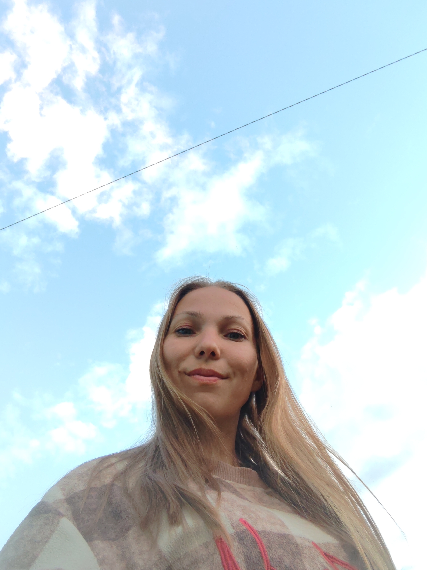 The image is a low-angle, upward shot of a young woman with long, light brown hair, taken outdoors against a bright blue sky with scattered white clouds. She is wearing a plaid sweater with cream, beige, and pink tones. A thin, dark diagonal line, possibly a wire, is visible in the upper portion of the sky. The woman is looking at the camera and smiling slightly, with a dimple visible on her left cheek. The scene is consistent with being in an outdoor urban or suburban setting, such as could be found in Чухлэм, Russia, where there are overhead wires and open sky.