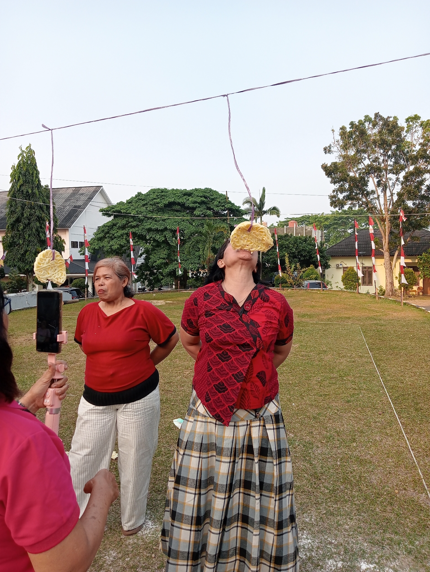 In an outdoor grassy area in Unknown, Unknown, two women are participating in a cracker-eating contest. A cracker is suspended from a string and held in front of the mouth of the woman on the right, who is actively trying to bite it. She is wearing a red patterned shirt and a plaid skirt. The woman on the left, wearing a red shirt and striped pants, observes the contest. A pink selfie stick with a phone attached is being held by another person, out of frame, likely recording the event. Indonesian flags are visible in the background, suggesting a national holiday or celebration. The sky is clear and bright.