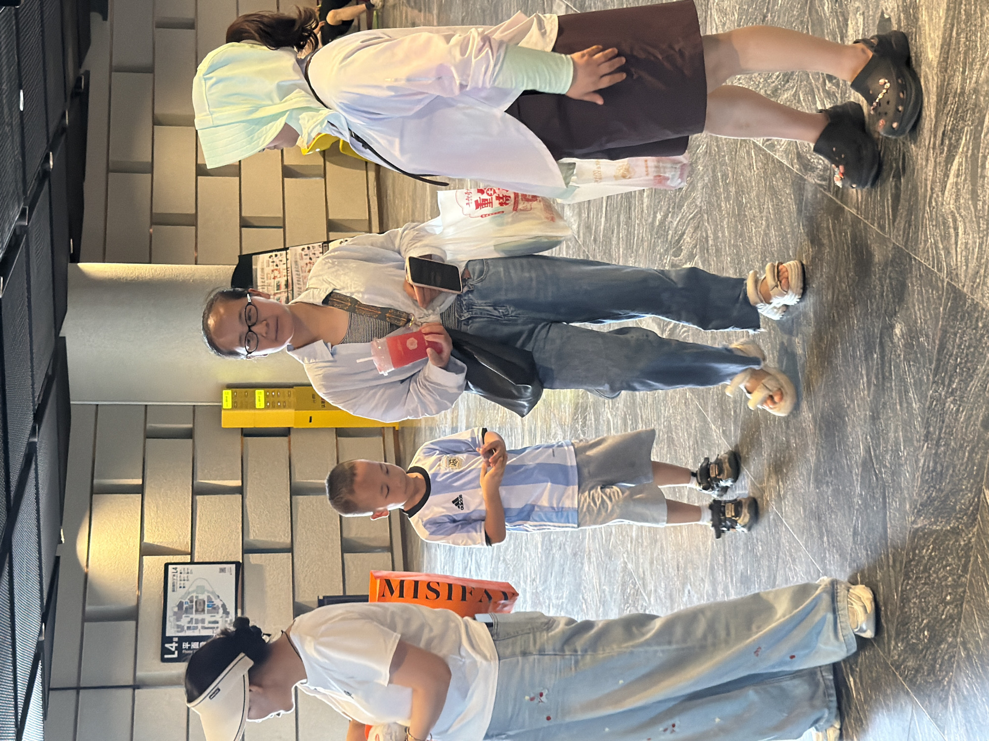 The image depicts a group of four people, two adults and two children, standing on a dark, polished floor that appears to be in a public indoor space, possibly a mall or an exhibition hall. The environment features a light-colored, brick-patterned wall behind the individuals.
On the left, a person wearing a white visor, a white t-shirt, and light blue wide-leg jeans stands facing away from the camera, holding an orange object with black text.
In the center, a boy, estimated to be around 6-8 years old, wearing an Argentina national football team jersey and gray shorts, looks down at his hands. To his right, a woman with glasses, wearing a light blue unbuttoned shirt over a striped top, blue jeans, and holding a pink drink and a phone, stands facing the viewer. She has a black shoulder bag.
On the far right, a person wearing a light green headscarf, a light purple long-sleeved shirt, and dark brown shorts stands with their back mostly to the camera, holding a white plastic bag.
The floor has a textured, marbled appearance. In the background, a yellow vertical display with multiple compartments is visible, along with a framed map on the wall. The lighting suggests an indoor environment with overhead lights casting shadows. The overall scene appears to be a casual gathering.