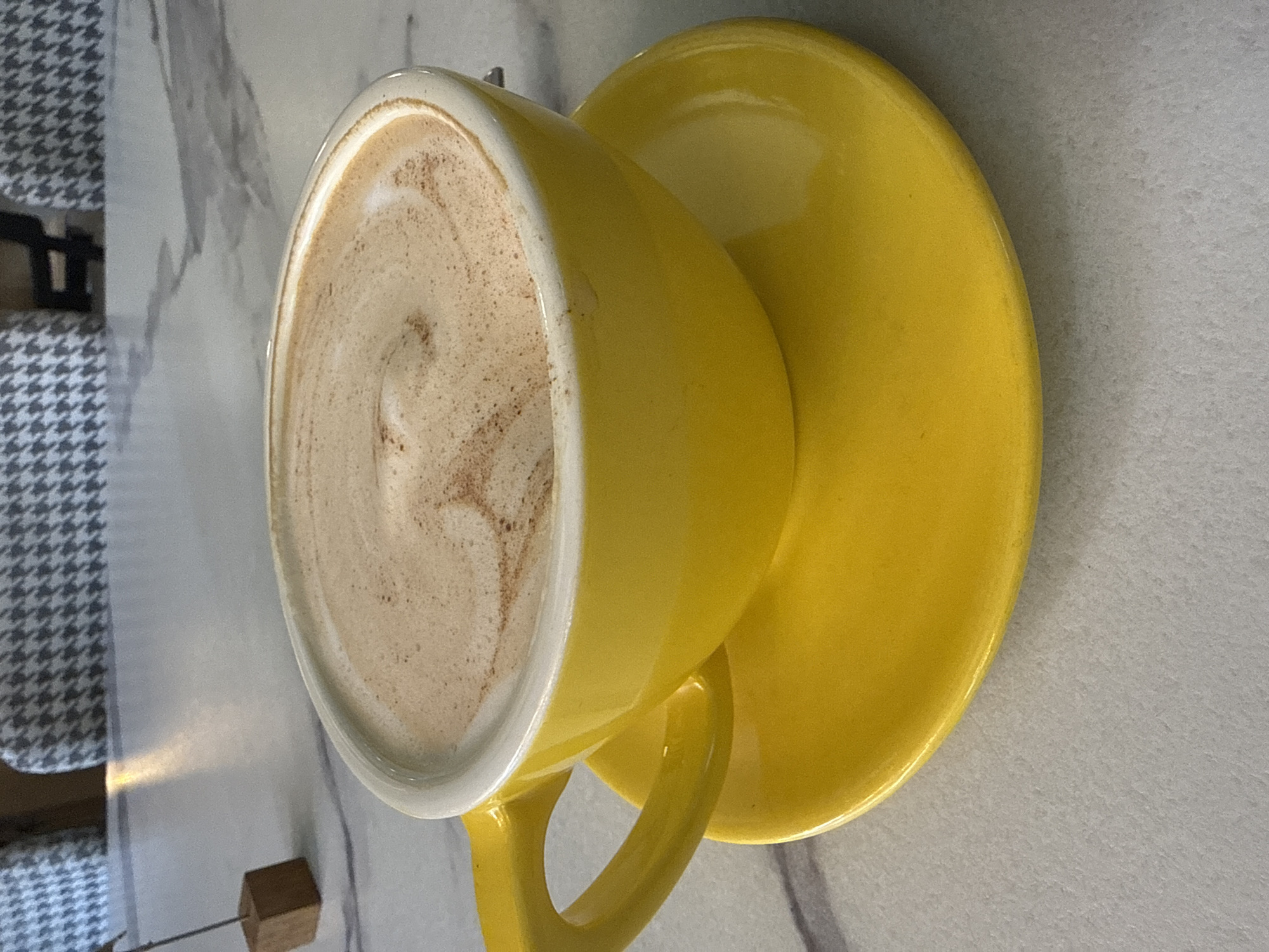 A close-up, eye-level shot shows a bright yellow coffee cup filled with a frothy beverage, possibly coffee or hot chocolate, topped with a swirl pattern and dusted with cinnamon. The cup and its matching saucer are on a light-colored, marbled surface. In the background, a small wooden block with a metallic rod sticking out is visible. Further in the background, two chairs with a grey and white houndstooth pattern are partially seen. The image is likely taken in a cafe or restaurant in Irkutsk, Russia, given the context of the request. There are no people visible in the image.