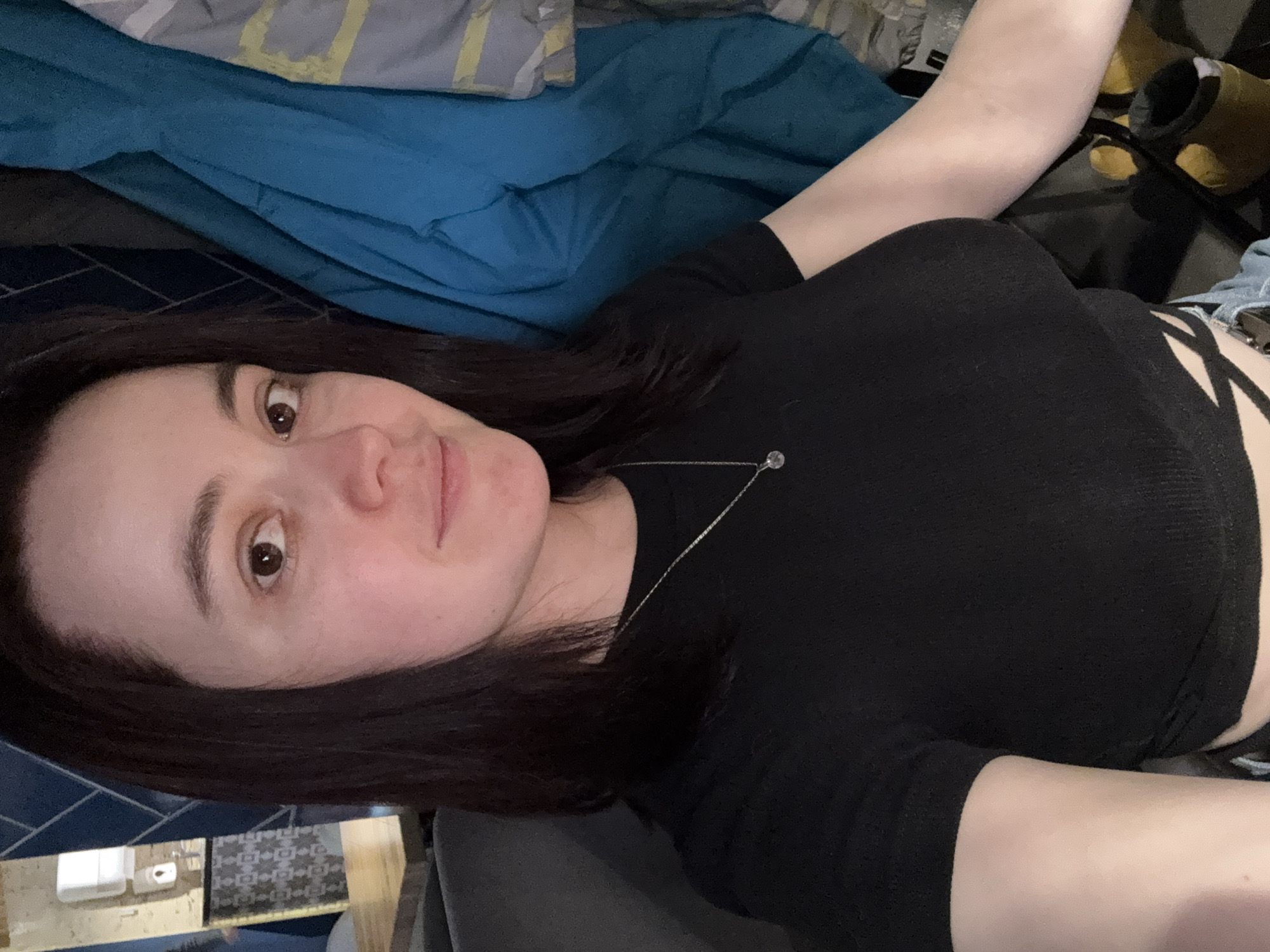 A woman with dark hair and fair skin lies on her back, wearing a black top and jeans. She is smiling at the camera. She is lying on bedding with blue and grey tones. The background includes a variety of objects including a black and yellow container. There is insufficient information to determine if the location is Izhevsk, Russia.