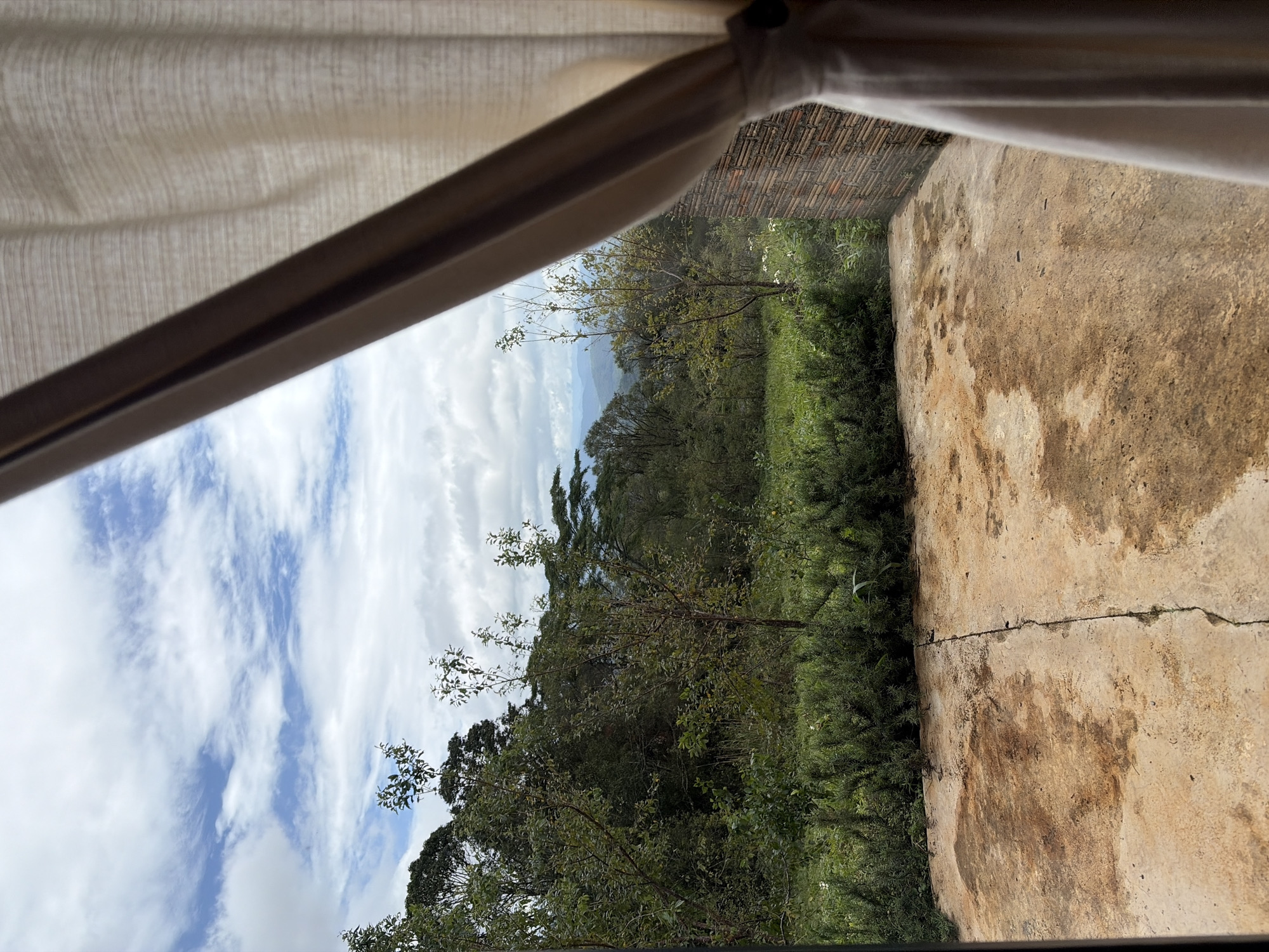 The image captures a view from an indoor space looking outwards through a partially drawn curtain. The scene depicts a natural landscape with lush green vegetation, trees, and distant mountains under a cloudy sky. The foreground shows a concrete patio with irregular brown stains. To the right, a portion of a brick wall is visible, and a cream-colored curtain hangs on the right side of the frame, obscuring part of the view. The environment suggests a location with a tropical or subtropical climate, possibly a resort or a private residence situated in a mountainous area. The overall impression is one of serene natural beauty and a connection to the outdoors. The image does not contain any people or explicit actions, focusing solely on the environment and architectural elements.