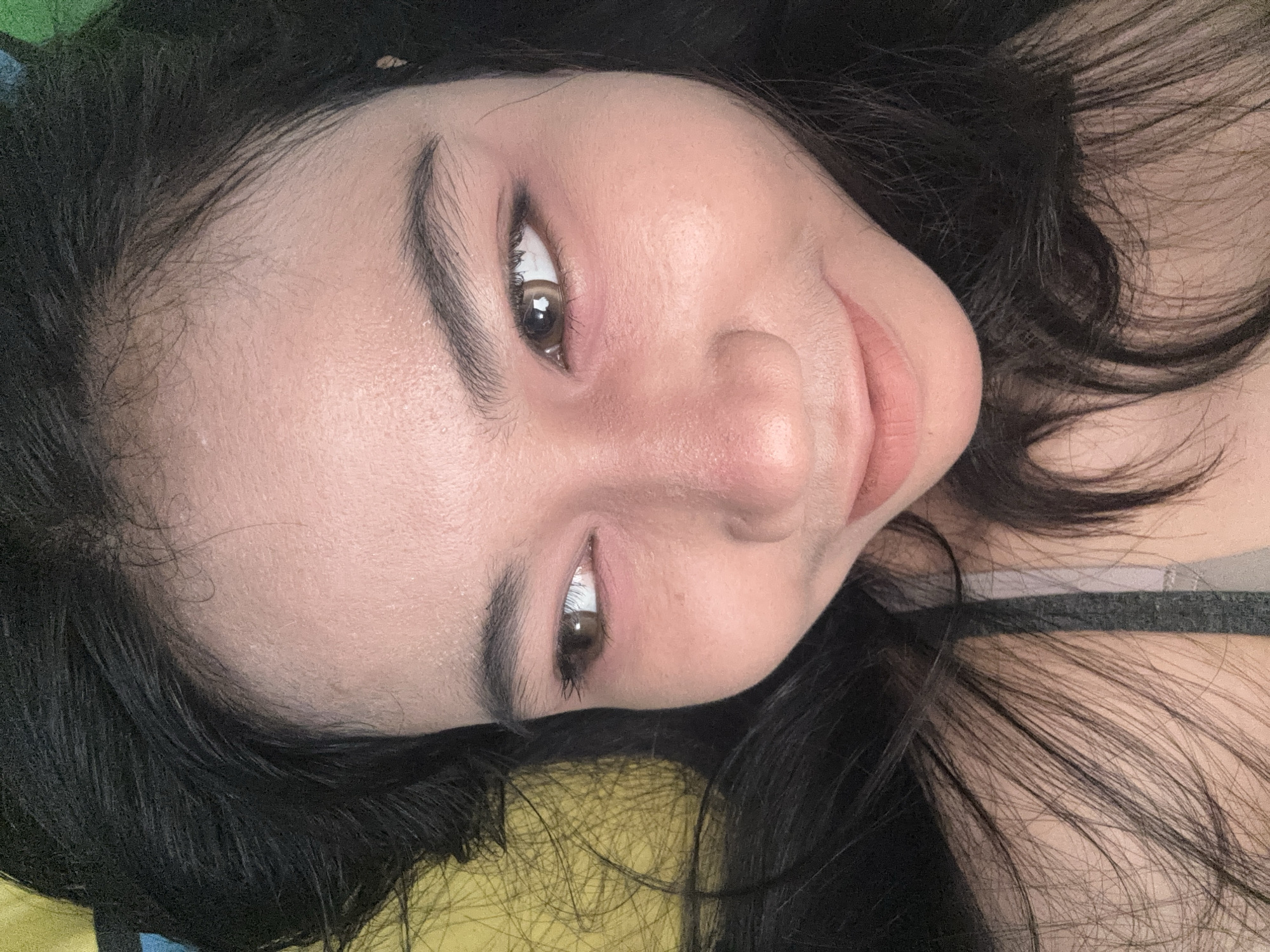 A close-up, tilted headshot of a woman with long, dark hair. She has light skin, brown eyes, and full lips. She is wearing eye makeup and possibly lip gloss. She appears to be lying down on a yellow and blue patterned surface. The lighting suggests an indoor environment. No specific details indicate a location in North Jakarta, Indonesia.