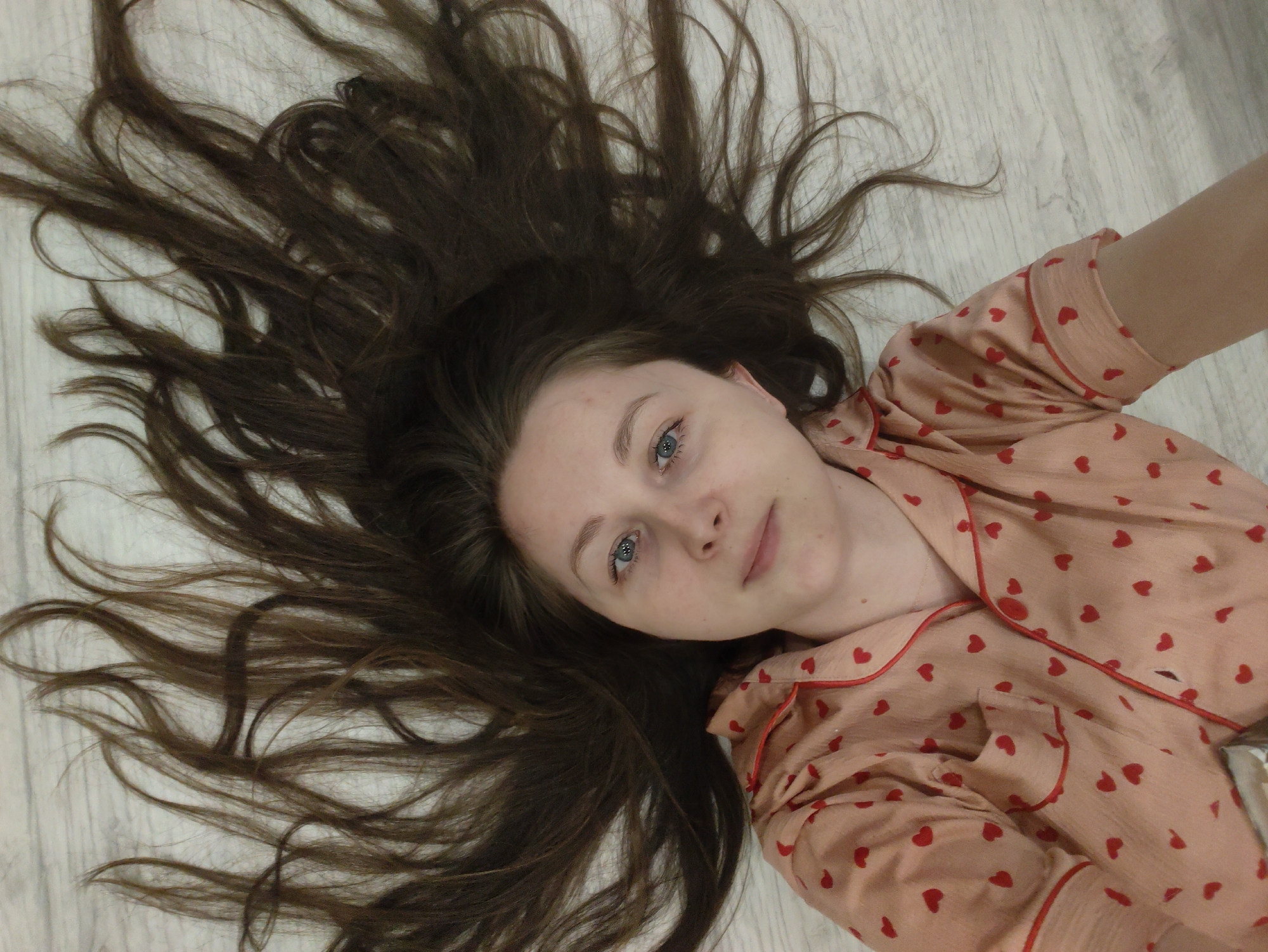 A young woman with long, flowing dark brown hair lies on a light-colored, wood-textured floor, looking directly up at the camera. Her hair is spread out dramatically around her head, creating a frame against the light background. She has fair skin and light blue eyes, with a neutral and serene expression, possibly a faint, relaxed smile. She is wearing a soft, light pink or peach-colored pajama top adorned with numerous small red heart patterns. The top features red piping along the lapels, collar, and cuffs, and appears to have a front pocket. The image is taken from an overhead perspective, capturing her upper body and the expansive spread of her hair on the floor. The setting appears to be indoors, likely a home, given the flooring and attire. There is no visible text.