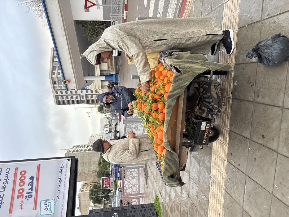 The image captures an outdoor street scene, likely in an urban area during daytime with an overcast sky. The overall impression is one of a typical market transaction taking place on a sidewalk.
In the foreground, three men are gathered around a wooden cart heavily laden with fresh oranges, many still bearing green leaves. The cart is covered partly by a patterned blanket. One man, presumably a customer, stands on the right side of the cart. He is wearing a light-colored, hooded djellaba (a traditional North African robe) and black sneakers, and is leaning over the oranges, seemingly selecting or inspecting them. His right hand is reaching towards the fruit.
Opposite him, on the other side of the cart, stands the vendor. He is dressed in a dark blue hooded jacket and dark pants, and also appears to be handling the oranges, possibly bagging them or arranging them. A third man, also wearing a light-colored djellaba and a dark beanie, stands further to the left, observing the interaction or waiting.
The sidewalk features tactile paving for visually impaired individuals. To the right of the cart, a black plastic garbage bag rests on the pavement. In the immediate background, behind the men and the cart, a decorative metal barrier or railing separates the sidewalk from the street.
The background reveals a bustling urban environment. Various multi-story buildings line the street, including one with blue and white elements that could be a commercial establishment or gas station. Several cars are visible on the street, and a red and white triangular traffic sign (likely a yield or warning sign) is prominent in the upper background. The sky is largely white and cloudy, suggesting an overcast day, and the pavement appears somewhat damp, perhaps from recent rain.
A large billboard dominates the far left of the image, displaying text in Arabic. The visible text reads:
- "حسبوها" (Hasabouha - "Calculate it" or "Consider it")
- "30000 درهم" (30,000 Dirhams)
- "مهداة." (Mouhdaat. - "Gifted." or "Offered.")
- "مع بنك أفريقيا، ديما" (Ma' Bank Afriqia, Dima - "With Bank Africa, always" or "forever")
- A logo featuring the words "كاين الحل" (Kayn L'Hal - "There is a solution") and possibly "التحل" (Al-Tahall) which might be a partial word or related to the slogan.
Below the cart, a label on a box reads "tio-toni".