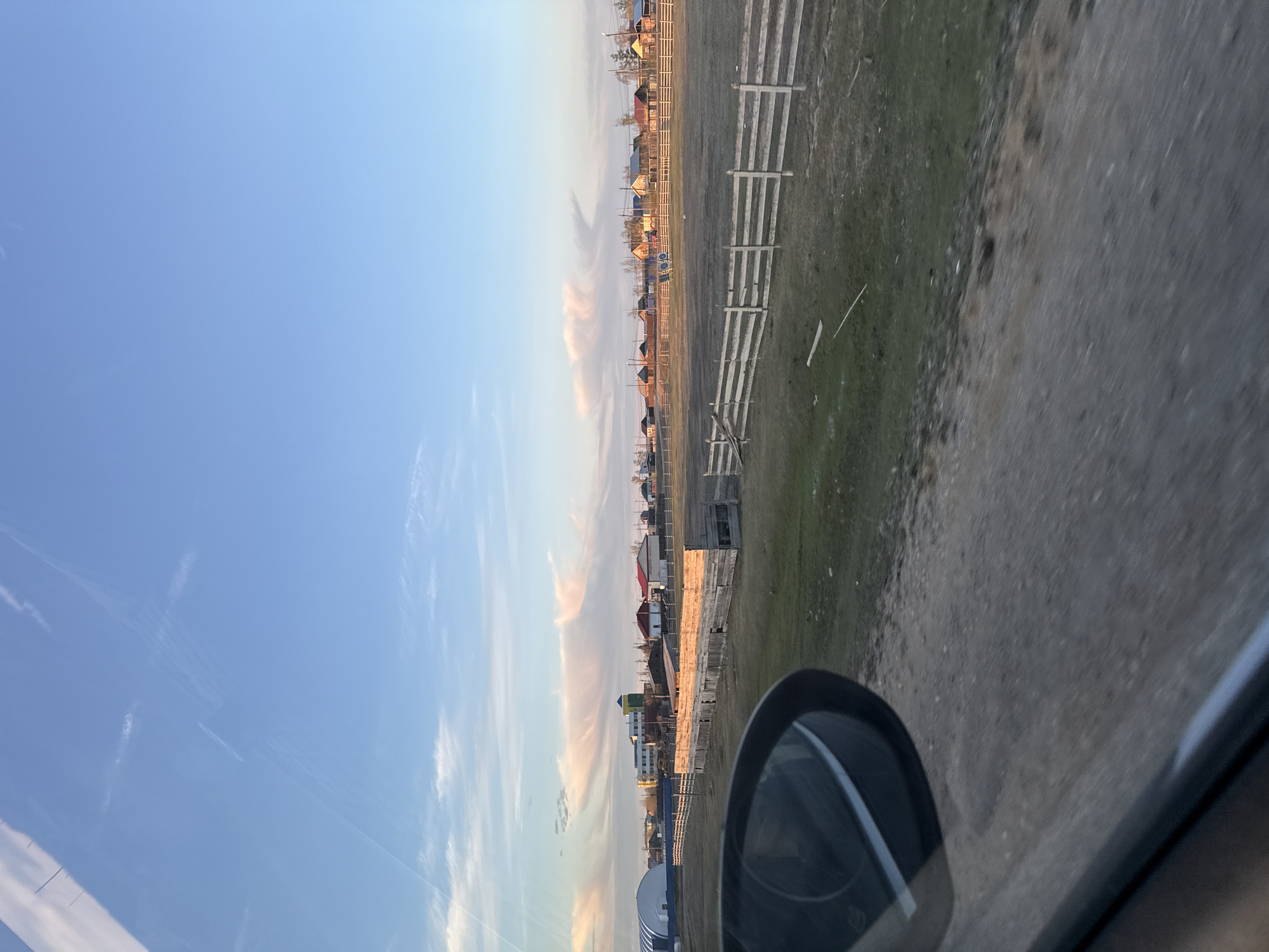 The image shows a view from inside a vehicle, likely a car, with the side mirror visible in the lower right. The landscape outside is mostly flat with a dirt road or field in the foreground. Beyond the road, there is a long, white fence running parallel to the road, with houses and buildings visible behind it. The sky is blue with wispy clouds. The location is likely Tulagino, Russia, based on the context provided. No people are visible. The scene suggests a rural or suburban setting.