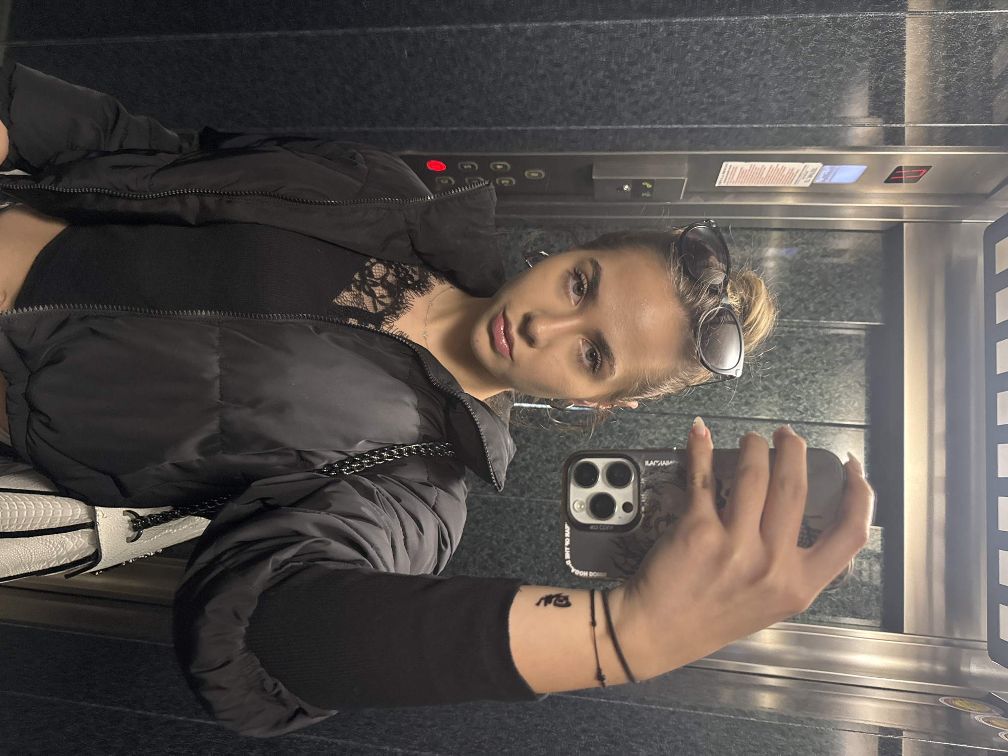 A young woman with blonde hair in a bun and sunglasses on her head takes a selfie in what appears to be an elevator with dark walls and metallic trim. She is wearing a black puffer jacket over a black top with lace detailing. She holds a smartphone in her right hand, which is visible in the reflection of the elevator's metallic wall. Her phone is dark-colored with a triple camera lens. A black bracelet is seen on her wrist along with what appears to be a small tattoo. Part of a white and textured handbag is seen to her left. The elevator buttons and floor indicator display are visible in the upper section of the photo. The location is not discernible.