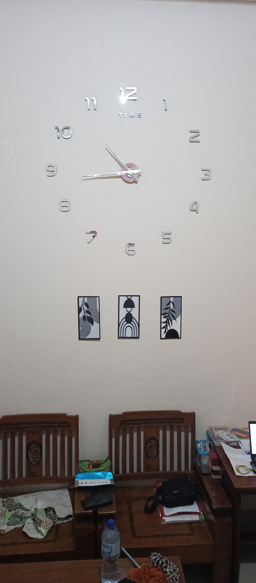 The image shows a wall with a large, modern clock and three framed pictures. The clock has silver, three-dimensional numbers attached to the wall, with a word "TIME" written below the number 12. The hands of the clock are silver and pink. The three framed pictures are rectangular and hung in a horizontal line. Each picture features a black and white abstract design with geometric shapes and nature-inspired elements. Below the pictures, there are two wooden chairs. On the left chair, there is a patterned cloth draped over it. On the right chair, there is a black bag, some papers, and a small stack of books. A bottle of water is visible on a lower surface in front of the right chair. The overall environment appears to be an indoor setting, possibly a living room or a common area.
The location is specified as the city of Arjasa, Indonesia.