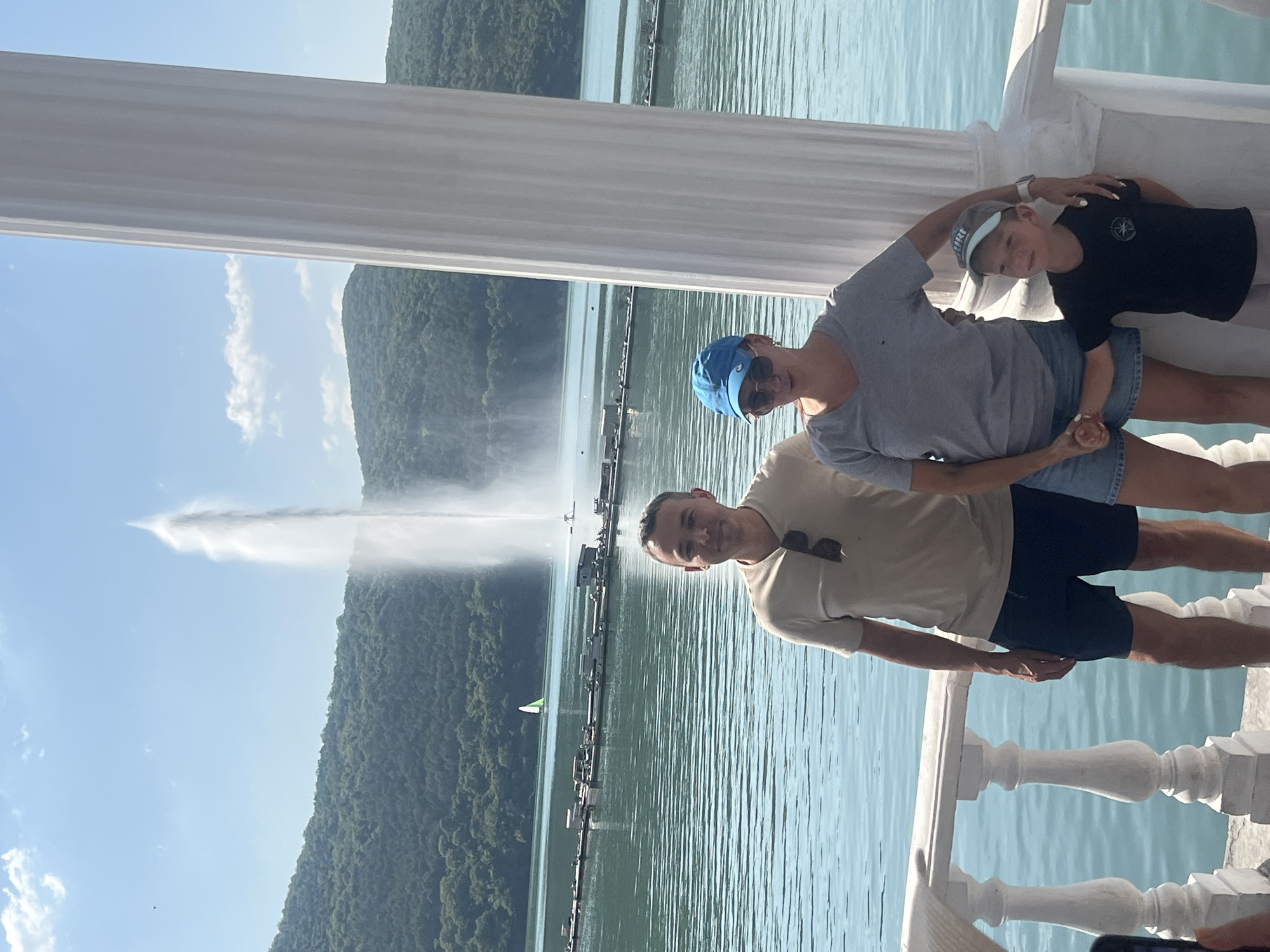 In the city of Abrau-Dyurso, Russia, a family of three is photographed on a sunny day. A man, woman, and boy stand in front of a large body of water, likely Lake Abrau, with a monumental fountain in the background. The man is wearing a beige t-shirt and dark shorts, while the woman sports a grey t-shirt and denim shorts, complemented by a blue baseball cap. The boy is dressed in a black t-shirt and grey shorts. They are positioned near a white ornate railing and a large white column, suggesting they are on a waterfront promenade or viewing area. The lake is calm and reflects the blue sky, which is dotted with a few white clouds. Lush green hills surround the lake, and a small green sailboat is visible in the distance. A long, floating structure with various compartments is present on the water, possibly a recreational area or part of the fountain's infrastructure. The overall scene conveys a sense of leisure and enjoyment of the natural landscape.