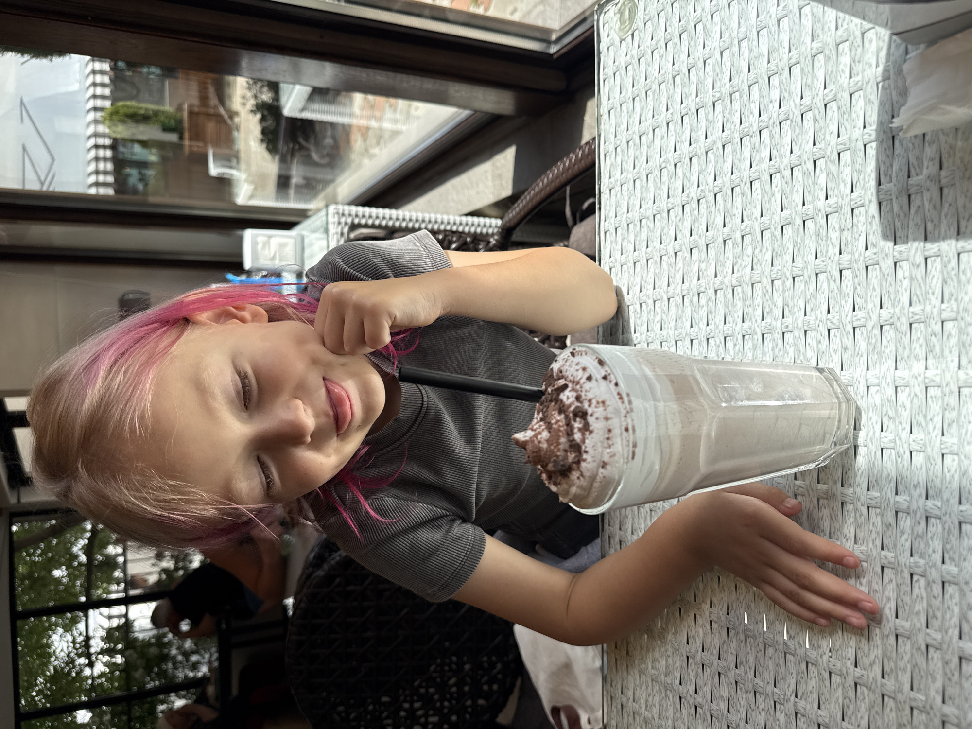 The image features a young person with light-colored hair, distinctly streaked with pink, sitting at a table. Their eyes are closed, and they have a relaxed expression with a slight smile. In front of them, on a light-colored, possibly white or off-white, woven textured table, is a tall glass containing a beverage. The drink, which appears to be a milkshake or a similar cold beverage, is topped with a generous swirl of whipped cream and dark sprinkles, likely chocolate. A black straw is inserted into the glass. The person is wearing a grey, horizontally ribbed short-sleeved top.
The setting appears to be a cafe or restaurant, possibly outdoors or near a large window, given the bright sunlight creating strong shadows on the table surface. Reflections in the background show architectural elements, including parts of buildings and possibly trees, suggesting an urban environment. The time of day is clearly daytime due to the bright light. The overall mood conveyed is one of contentment and enjoyment. There is no visible text in the image.