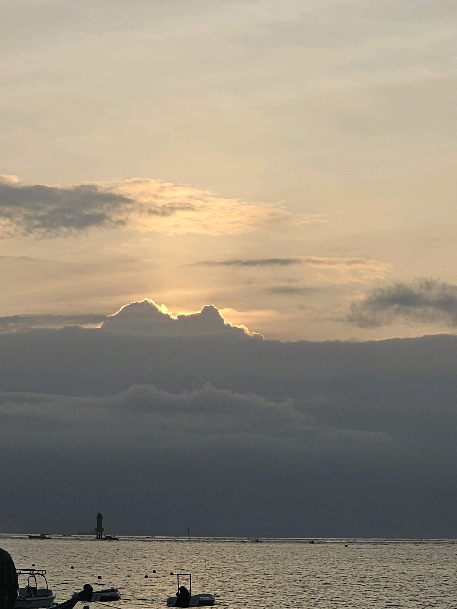 The image depicts a serene seascape at sunset or sunrise, likely along the coast of Denpasar, Indonesia. The sky is filled with a mix of soft, pale yellow and grey clouds, with the sun's light brilliantly illuminating the edges of some clouds, creating a halo effect. Below the sky, a vast expanse of calm water reflects the soft light. In the distance, a slender lighthouse stands on the horizon, a solitary sentinel against the muted sky. Several small boats are scattered across the water, some docked near the shore, others appearing to be out at sea. The overall atmosphere is one of tranquility and natural beauty, characteristic of coastal scenes in Denpasar.