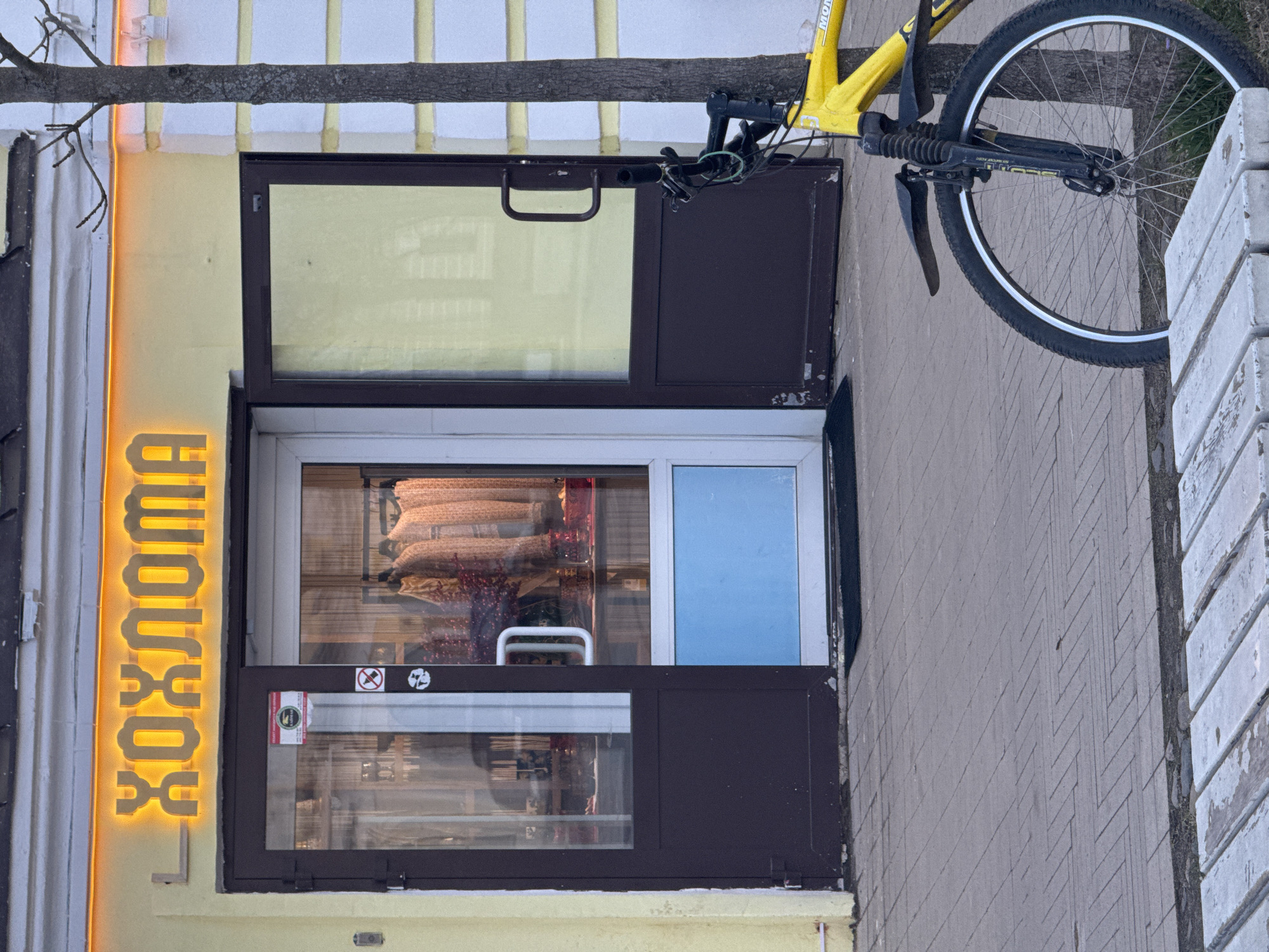 The image shows the exterior of a small storefront in Novo-Nikolskoye, Russia. The sign "ХОХЛОМА" is illuminated vertically on the left side of the storefront. The storefront has a multi-paned window with a dark brown frame. The top pane is open like an awning, while a lower pane displays items inside. A yellow bicycle is partially visible on the right side of the frame, with the front wheel appearing most prominently. The building's facade is a combination of light yellow and gray materials. The sidewalk is made of gray brick or concrete. A portion of a white painted fence is visible in the background on the right.