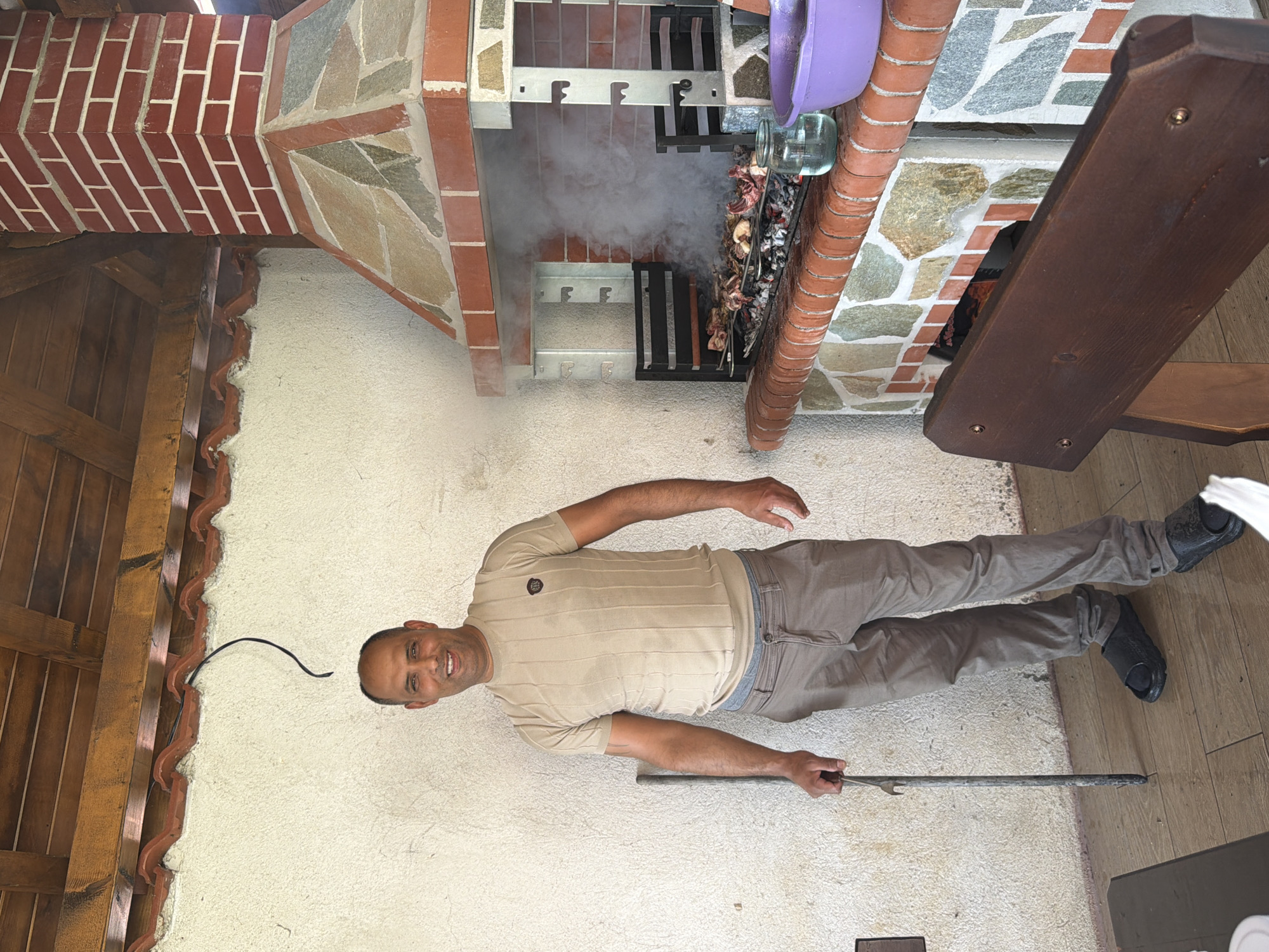 This image, captured in Sinitovo, Bulgaria, shows a male subject standing on a tiled outdoor patio next to a large, active barbecue grill under a wooden-beamed roof. The man, appearing to be in his 40s or 50s with short dark hair and a light complexion, is smiling broadly and looking directly at the camera. He is dressed in a beige short-sleeved, ribbed knit shirt with a dark emblem on the left chest, light brown casual trousers, and black slip-on sandals. His right hand grasps a long, slender metal rod, likely a barbecue tool.
To the man's right, a permanent barbecue structure dominates the frame. Its lower section features a mosaic of various colored natural stones and red tile accents, topped by a red brick countertop. Actively cooking meat, consisting of multiple pieces, is visible on a charcoal grill within the barbecue, producing a significant amount of white smoke. The grill system includes adjustable metal racks on both sides. Above the cooking area, a red brick chimney with a decorative stone hood extends upwards towards the roof.
On the barbecue countertop, a clear glass jar containing a clear liquid and a purple plastic bowl are present. The background behind the man and to the left of the barbecue is an off-white, textured stucco wall, showing minor cracks. A black electrical cable is visibly hanging from the wooden ceiling rafters against this wall. In the immediate foreground, to the lower right, the dark brown wooden backrest of a bench or chair is visible. The floor is covered with large, rectangular, light brown tiled planks. The overall scene suggests an outdoor cooking and leisure setting during daylight hours.