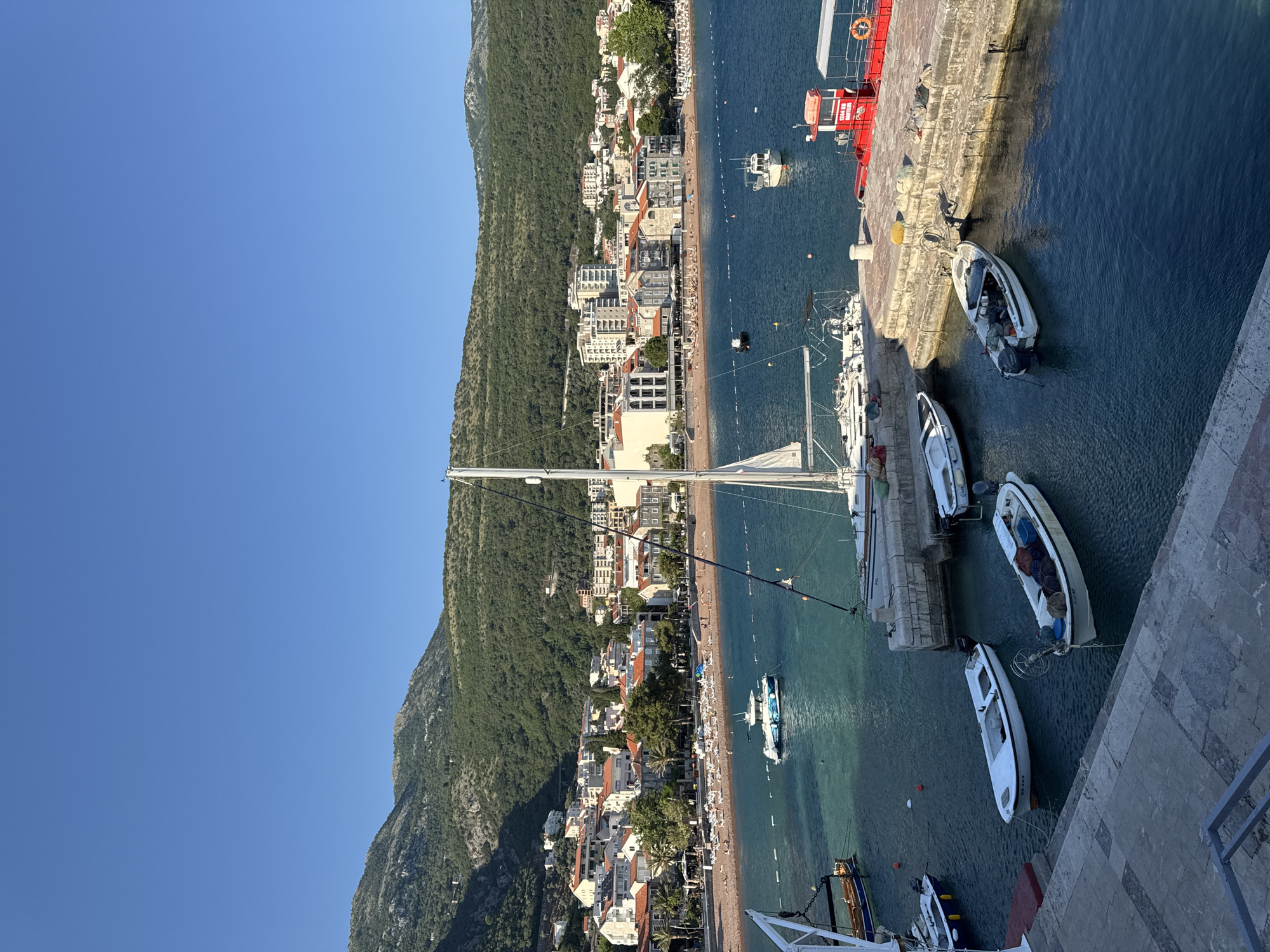 The image shows an elevated perspective of a coastal scene in Petrovac centar, Montenegro. A coastline featuring a densely populated town with buildings, trees, and a sandy beach borders a clear blue sea. A green mountain rises sharply behind the town. Numerous boats are docked along a concrete pier at the edge of the town, with other boats scattered in the water along the beach. People are visible on the beach. A sailboat's mast is prominent in the foreground. The sky is clear and blue.