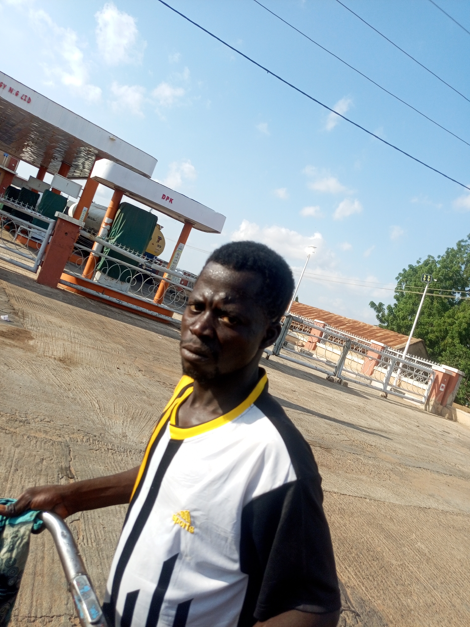 A man, wearing a white, black, and yellow striped athletic shirt, is standing outside a gas station. The gas station has an orange and white canopy with "DPK" visible on the side. The man's right hand is holding onto the handle of a bicycle. He is looking towards the camera with a serious expression. The ground is paved with concrete, and there are some scattered pieces of debris. In the background, there are trees, power lines, and a building with a rusted roof. The sky is clear with a few white clouds. The overall lighting suggests it is daytime. The man's attire and the general atmosphere indicate a likely urban or semi-urban setting in Gombe, Nigeria.