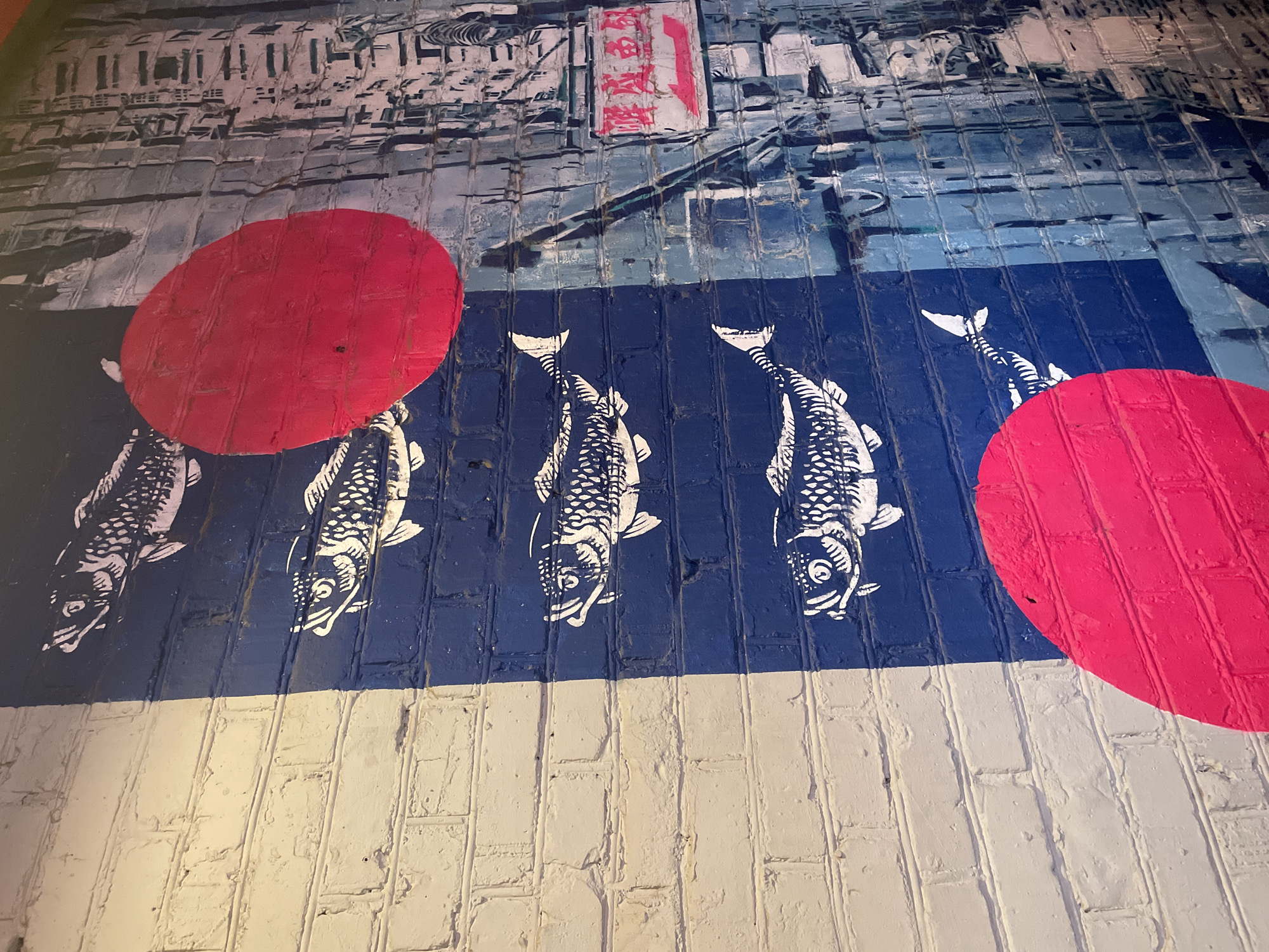 The image shows a brick wall with a mural. The top third of the wall is painted in shades of blue and white, depicting a cityscape with structures and a sign in a language that is not clearly legible. Below that, a horizontal blue stripe is painted with white koi fish swimming in one direction. Two large red circles are painted over the fish and background elements. The bottom third of the wall is painted white. The brickwork itself is visible throughout the painting.