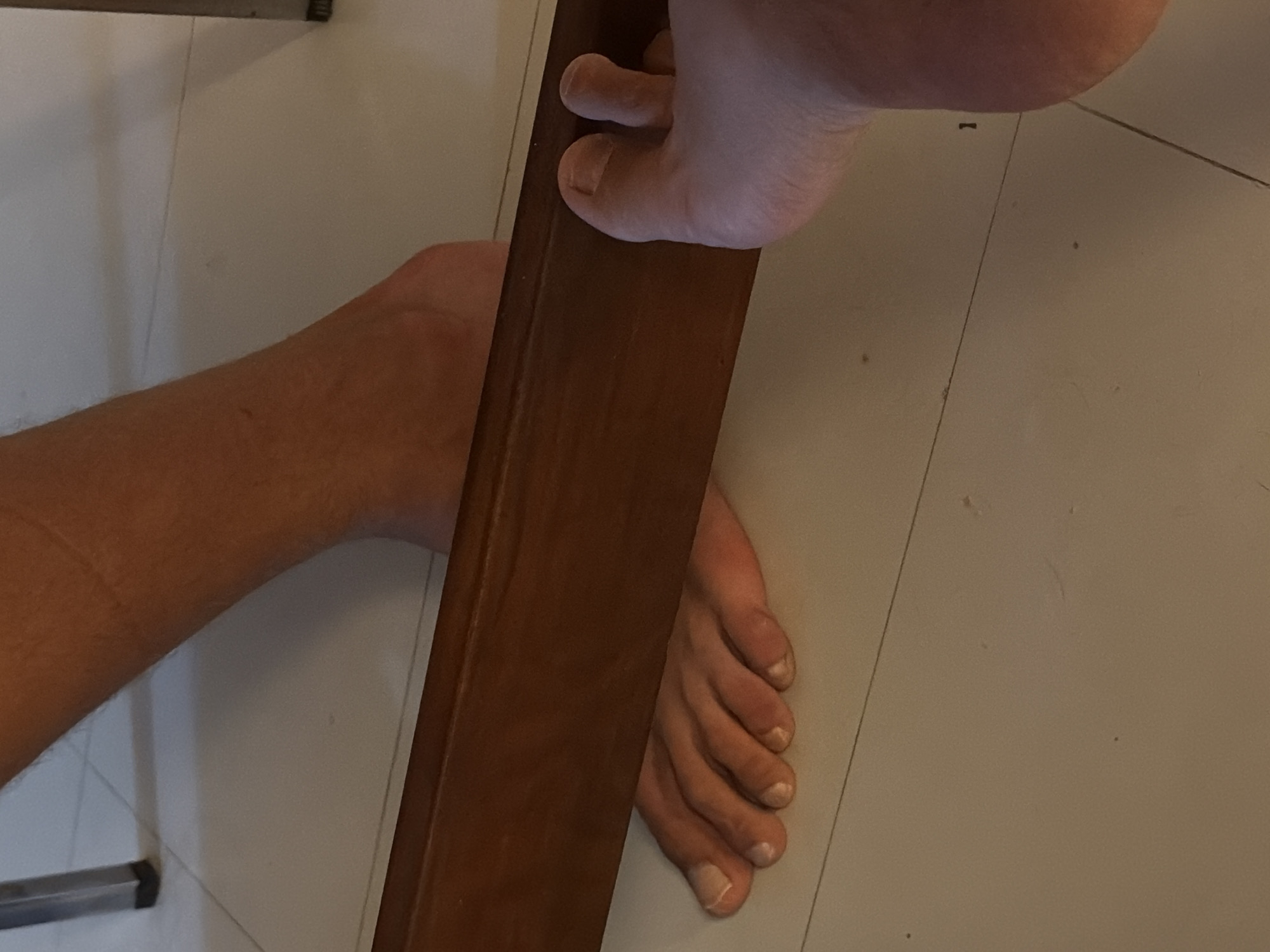 An overhead close-up shot captures two bare feet positioned on a light-colored tiled floor, separated by a diagonal dark brown wooden object. On the left, the arch and lower calf of one foot are visible, resting casually on the floor. On the right, the toes and forefoot of another foot are prominently displayed, with the toes curled over and gripping the top edge of the wooden object. The toenails on both feet are visible and appear natural. The wooden object is rectangular and smooth, possibly a furniture leg or beam. The tiled floor has visible grout lines. In the bottom left, a metallic furniture leg with a black cap is partially visible. The scene is indoors with ambient lighting, casting soft shadows. The activity appears to be someone resting or playfully interacting with furniture. No text is visible in the image. The general location context is Chang Phueak, Thailand.