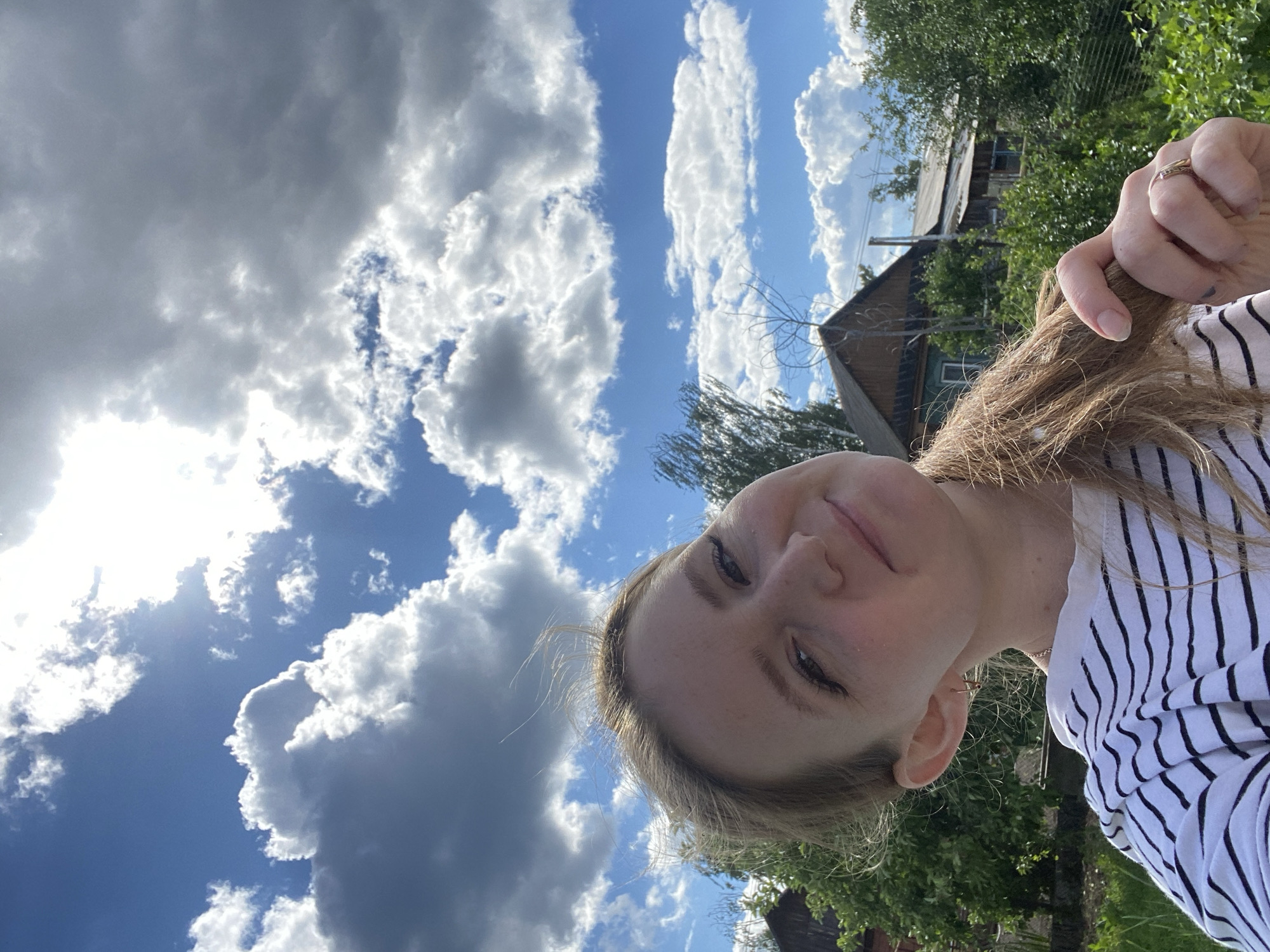 A young woman with blonde hair, wearing a black and white striped shirt, takes a selfie outdoors. She is looking up towards the sky, which is filled with dramatic white and grey clouds against a bright blue background. The sun is shining through the clouds, creating bright spots and shadows. In the background, there are trees, lush green foliage, and the roofs of wooden houses, suggesting a rural or suburban setting. A ring is visible on her finger as she plays with her hair. The overall impression is of a bright, sunny day in what appears to be a village or countryside environment.